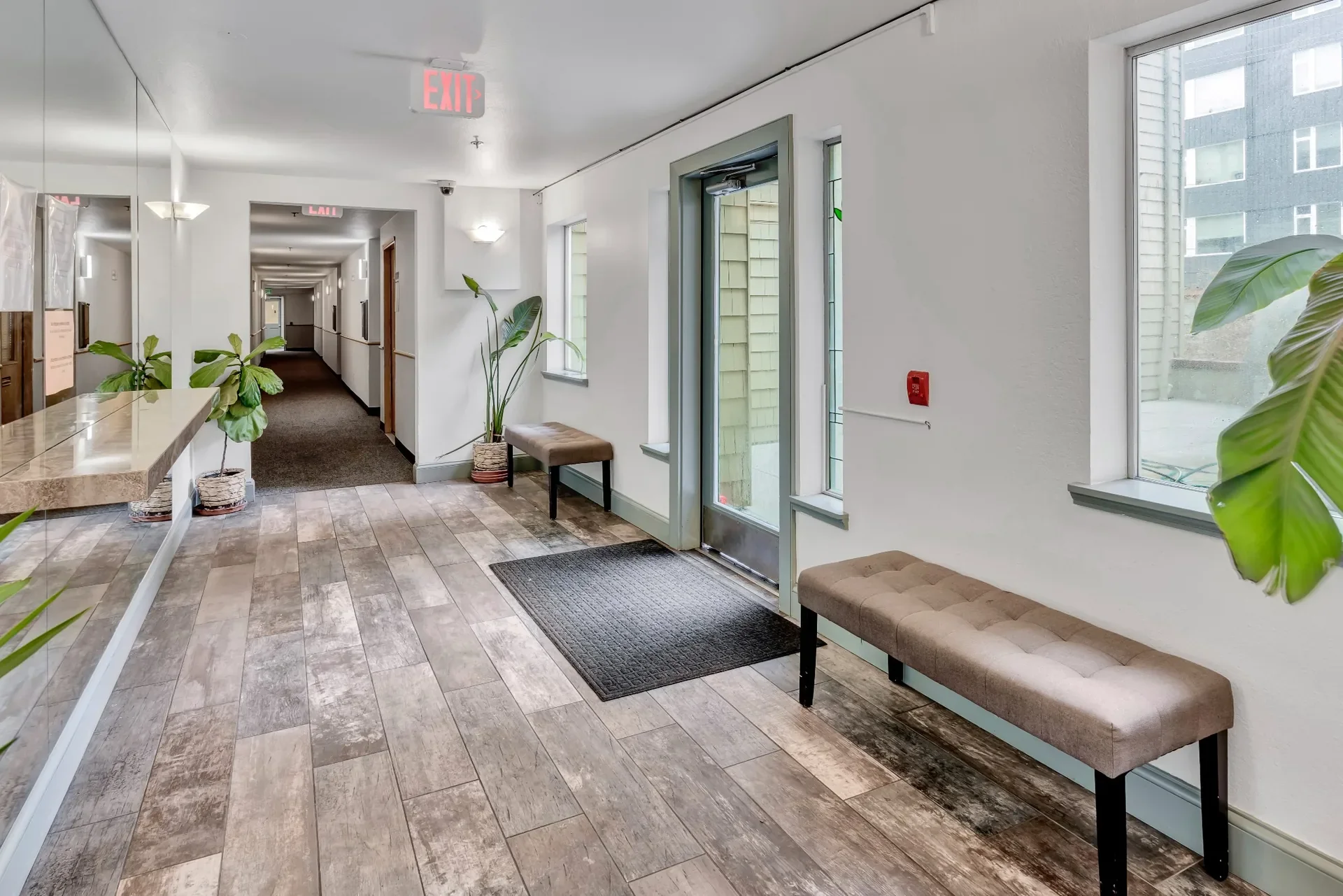 Interior of a building lobby with windows, benches, potted plants, and an exit door visible in the background.