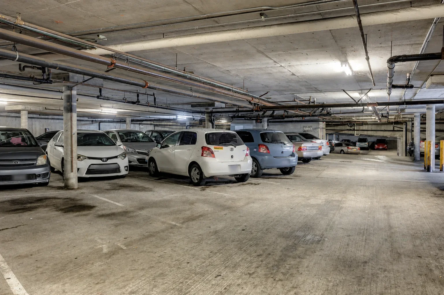 Underground parking garage with several parked cars, concrete ceilings, and exposed piping.