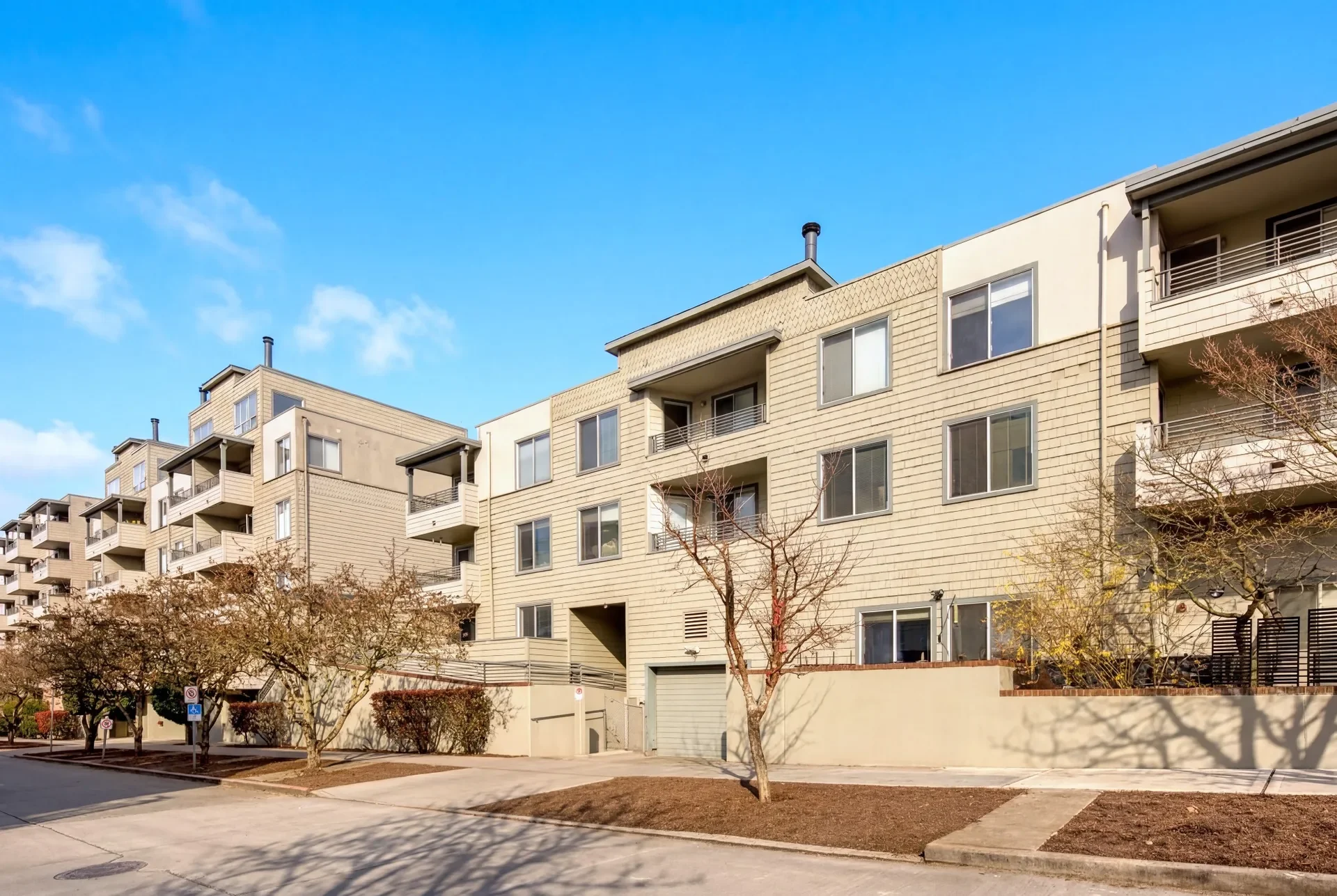 A multi-story residential building under a clear blue sky. The building has beige siding, multiple balconies, and several windows. There are leafless trees in front of the building, and a sidewalk and parking area with a few vehicles.