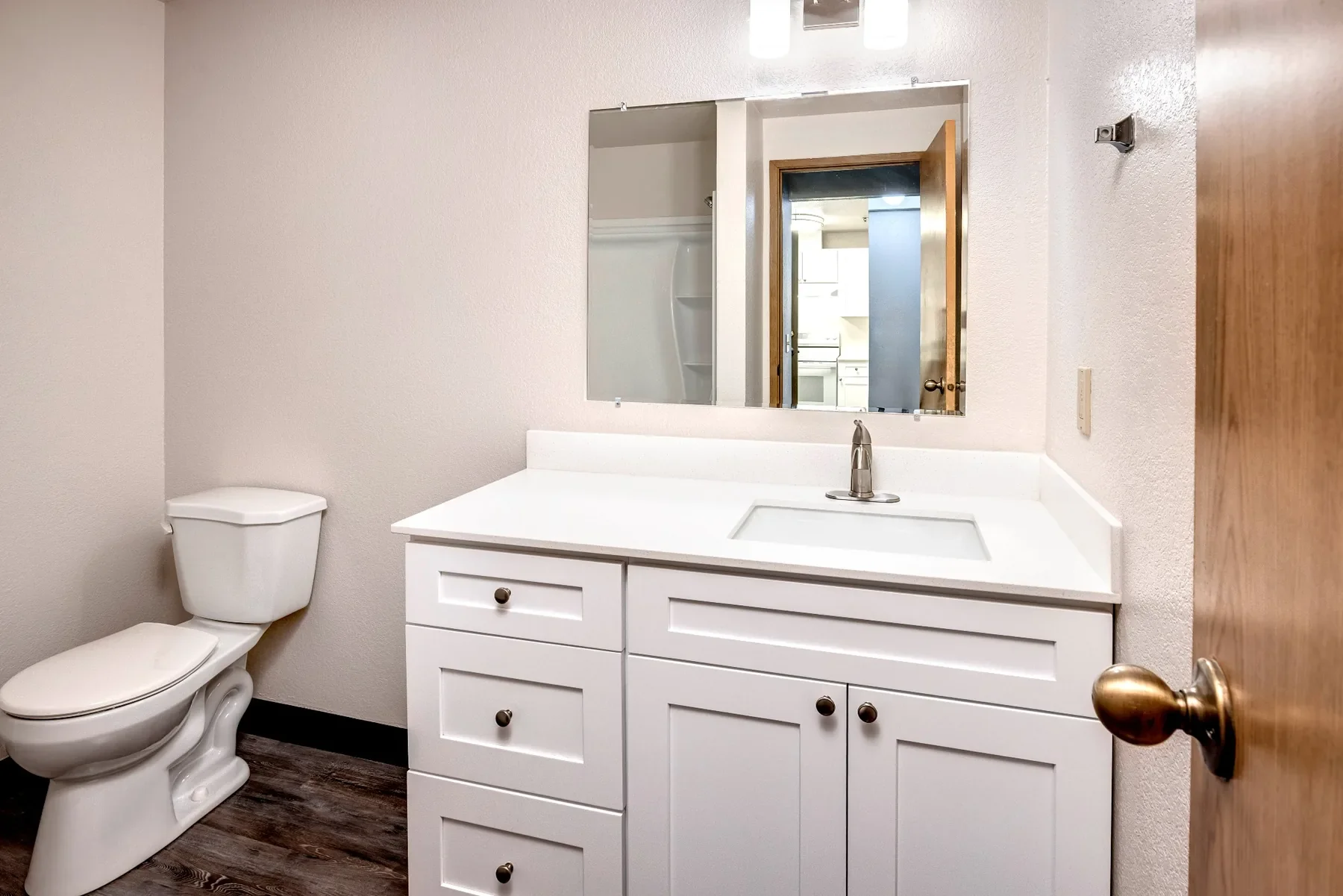 A bathroom with a white toilet on the left, a white vanity with a sink in the middle, a mirror above it, and a door with a brown finish on the right. The floor has a dark wood pattern, and the walls are painted light beige. Visible in the mirror's re