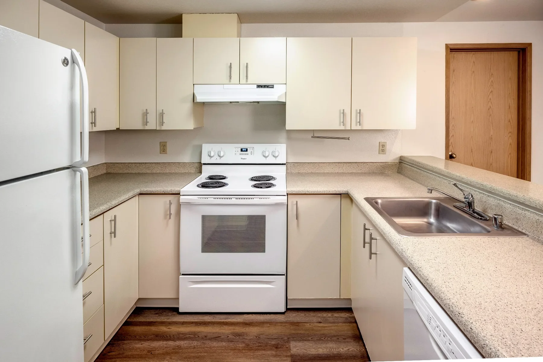 Kitchen with white refrigerator, white stove, microwave, beige cabinets, stainless steel sink, and beige countertops. Wooden floor and a wooden door are visible.
