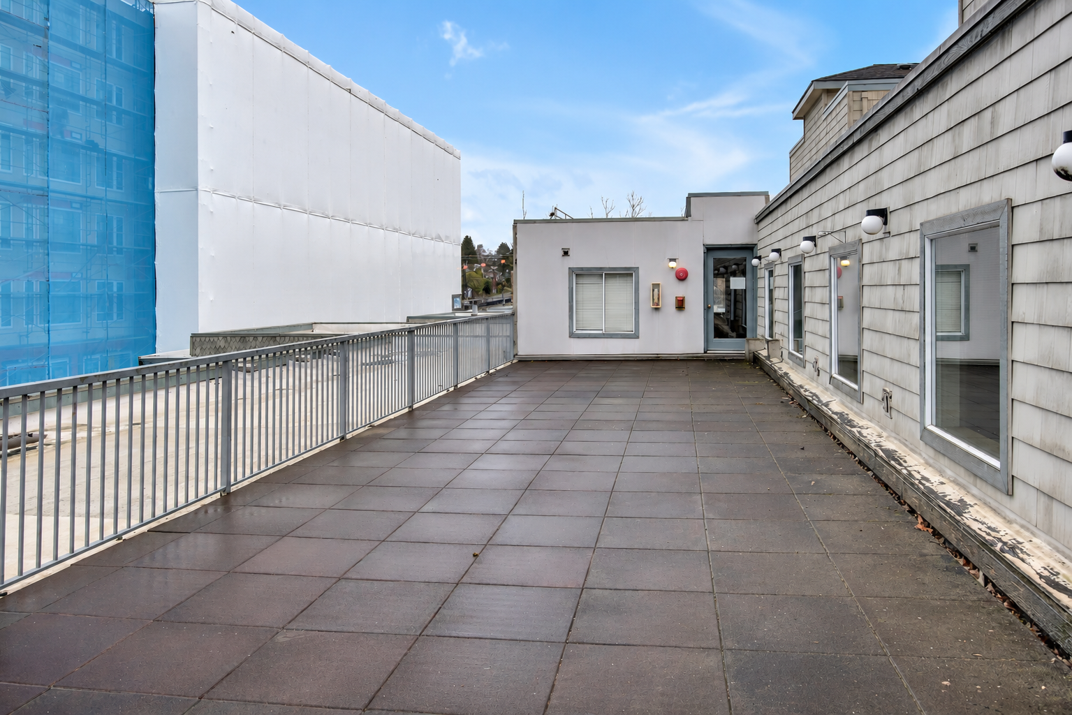 Empty rooftop patio with brown tiles, metal railing, and building walls with windows and exterior lights.