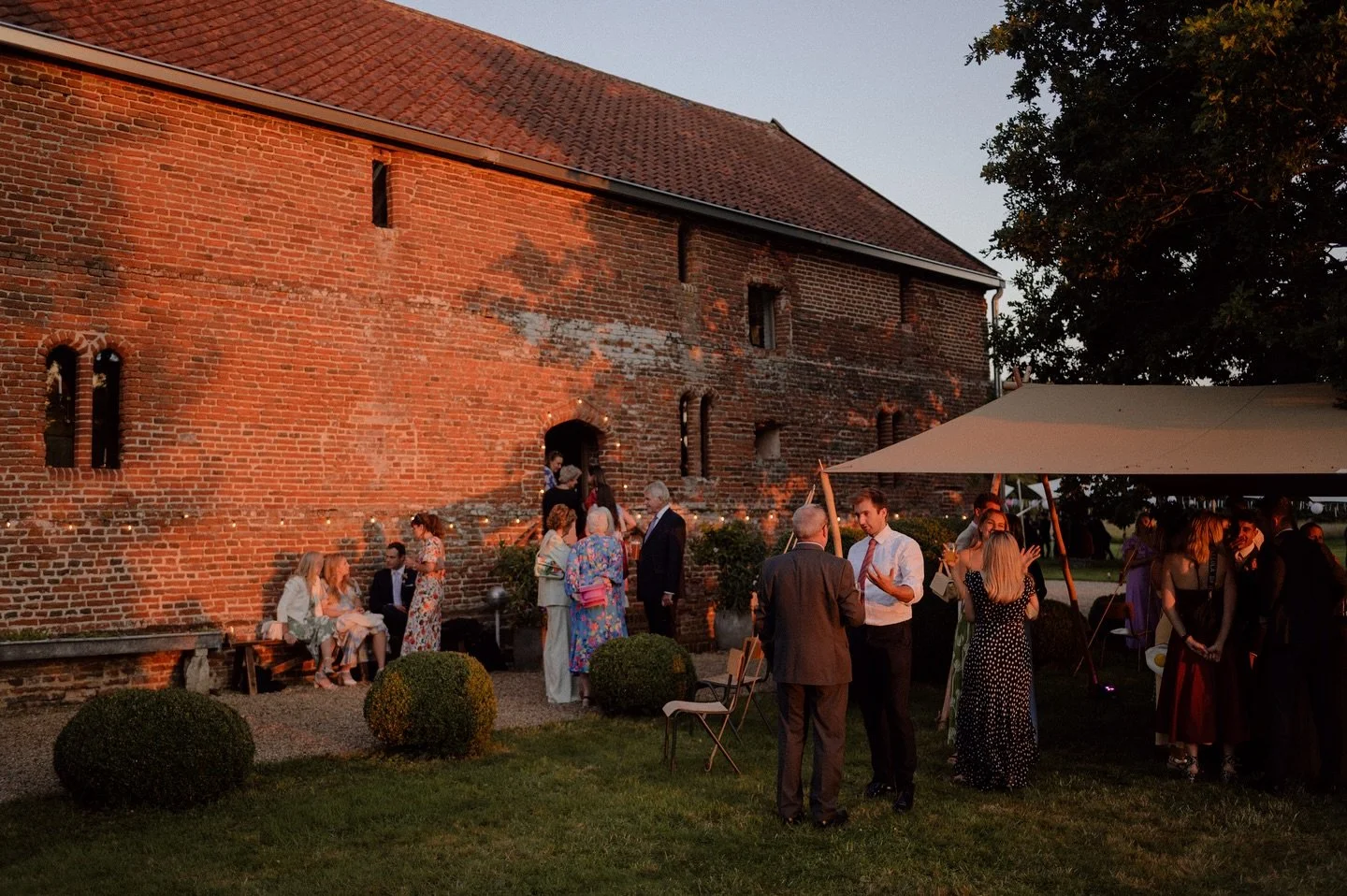 People gathered outside under a tent for an evening event near a red brick building with small windows, during sunset.