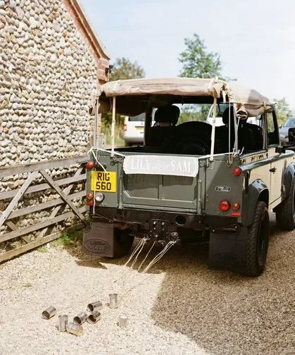 A vintage green Land Rover parked next to a stone building with a wooden fence. The vehicle has a soft top with a sign that reads 'Lilly & Sam' on the back and a yellow license plate with 'RIG 560'. Some metal pipes are scattered on the gravel ground nearby.