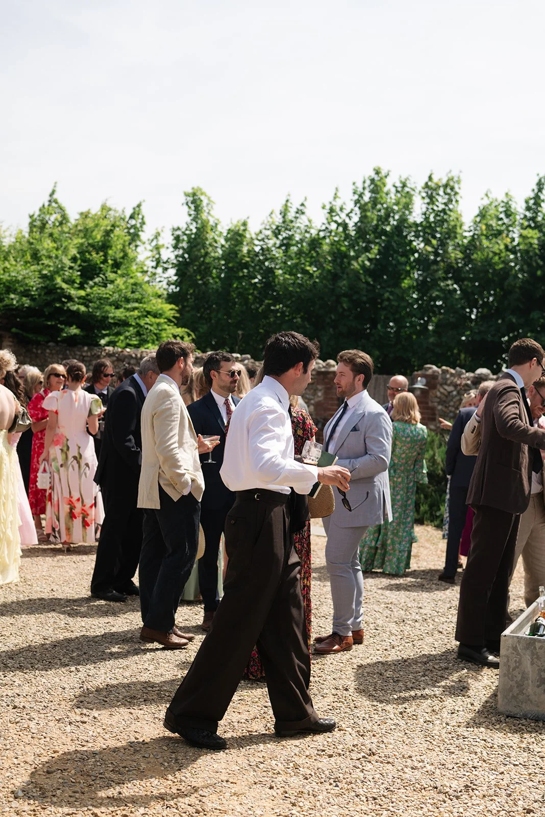 People dressed in formal attire gather outdoors, socializing at a sunny event with green trees and a stone wall in the background.