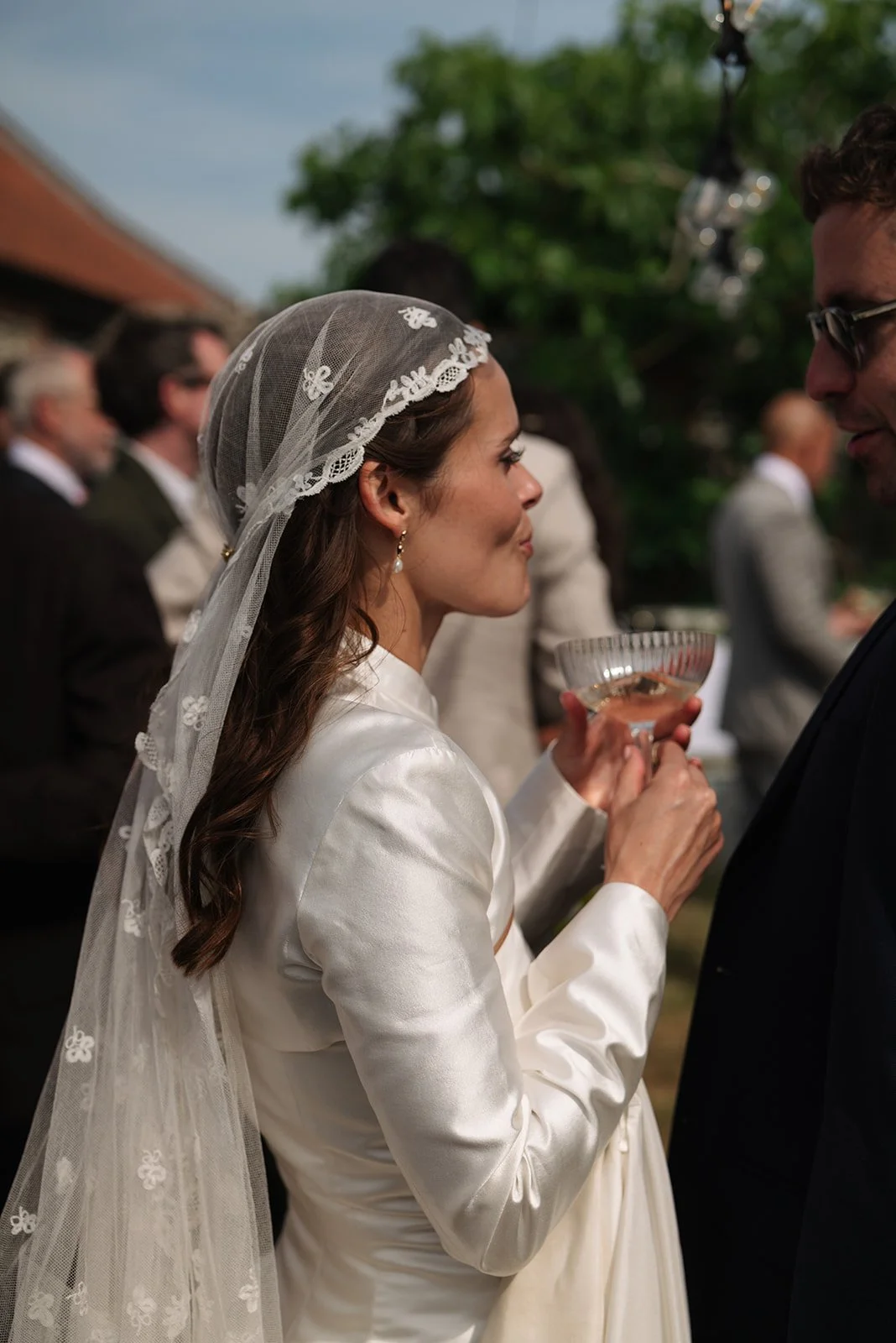 A bride with long brown hair holding a glass of champagne or cocktail during her wedding ceremony, wearing a white satin gown and lace veil, standing face-to-face with a groom in a dark suit, outdoor setting with guests in the background.