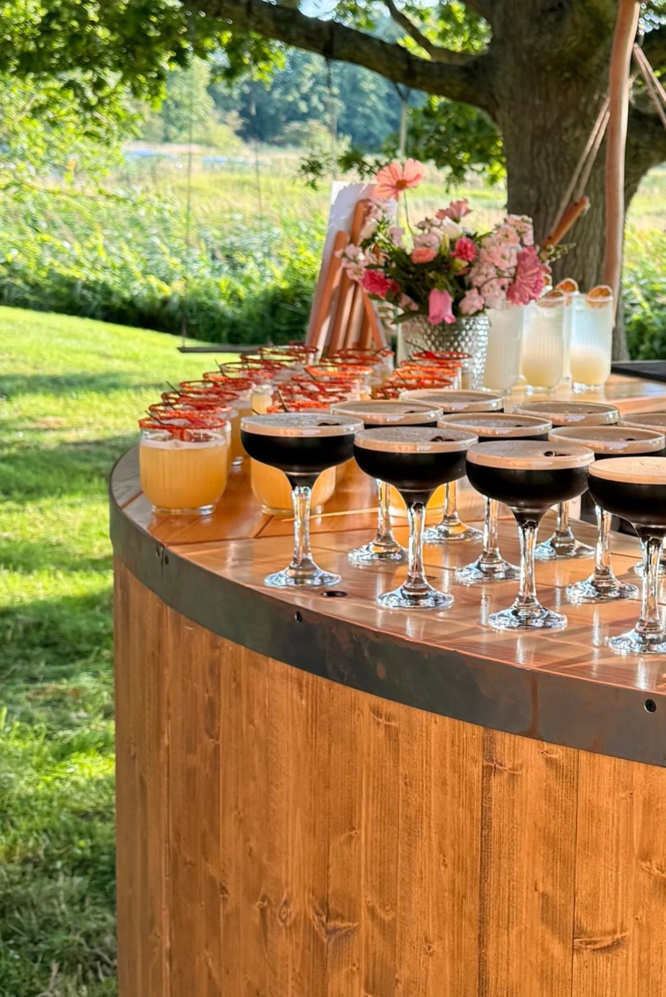 A wooden bar table set outdoors under a large tree with green leaves. There are glasses of different drinks, some topped with blueberries, and a floral centerpiece with pink and white flowers.