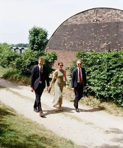 Three people walk on a dirt path with greenery and a large brick wall in the background.