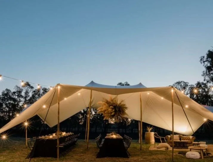 Outdoor event space with a large tent canopy, string lights, tables, and chairs at dusk.