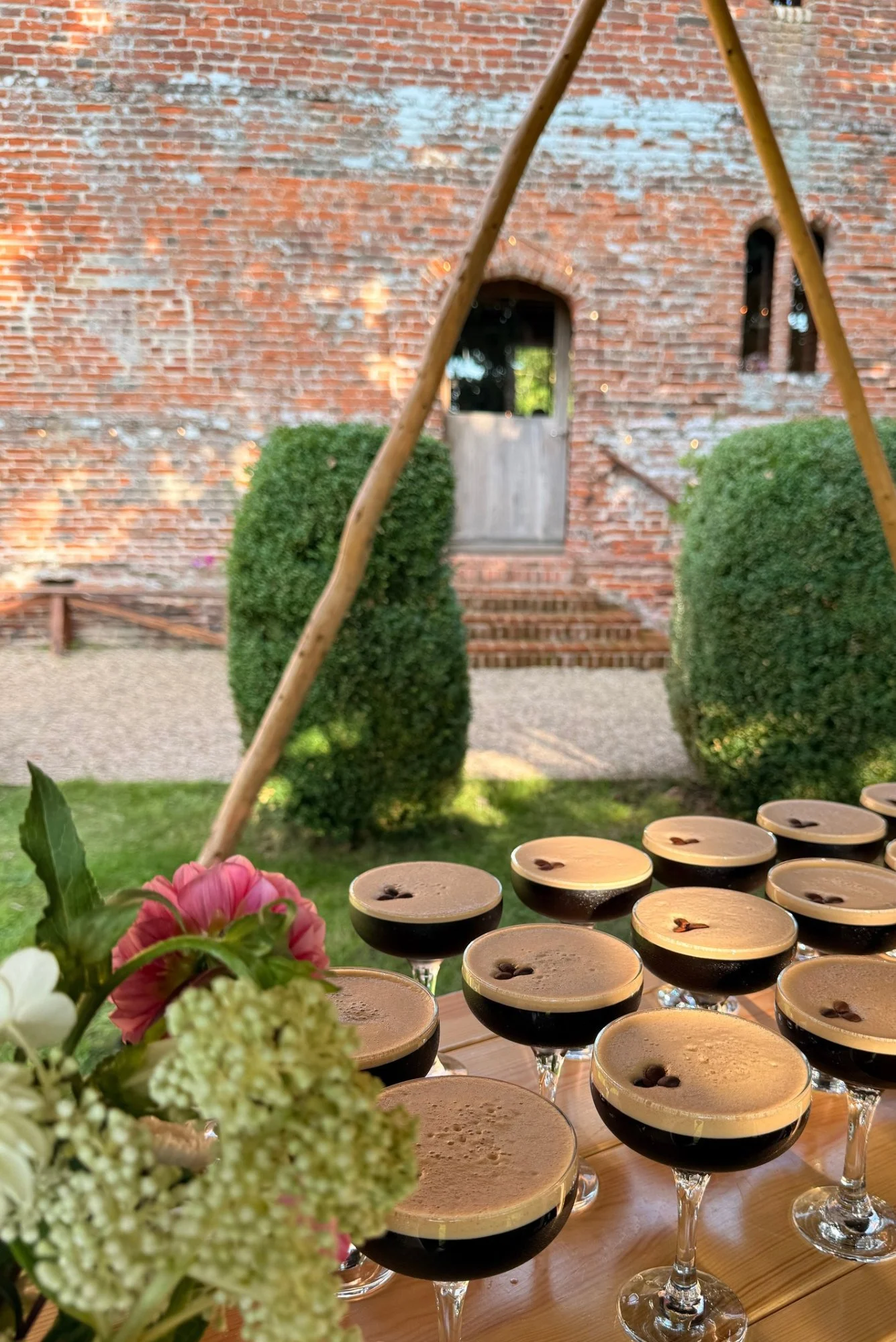 Multiple glasses of dessert with a chocolate top, decorated with a small flower in each glass, set on a wooden table. In the foreground, there are colorful flowers. The background features a brick wall with a small arched door and window, surrounded by greenery.