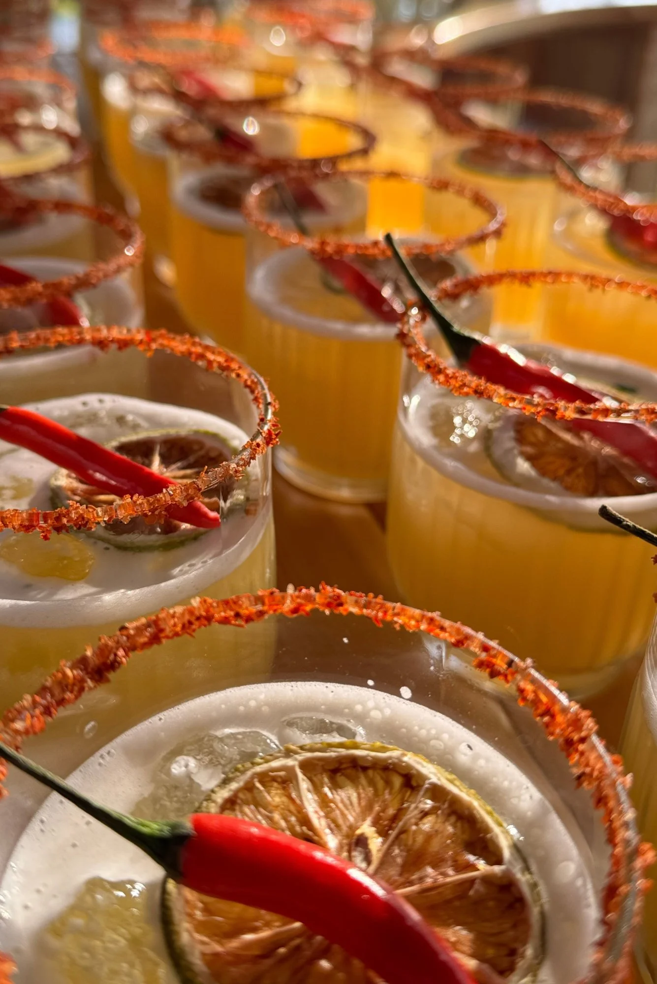 Close-up of glasses with yellow drinks garnished with lime slices and salted rimmed lids, with red chili peppers, some garnished with dried chili flakes.