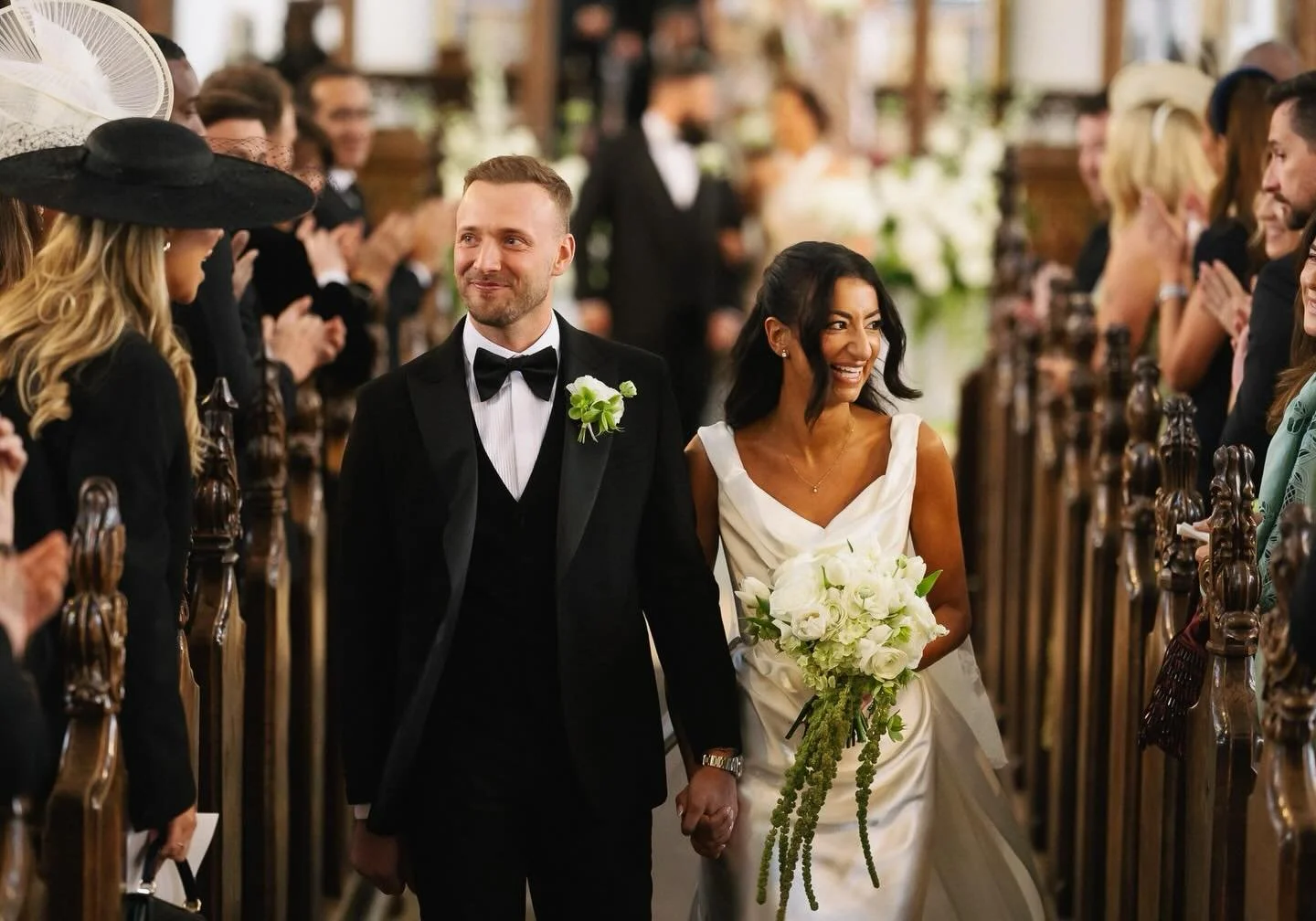 A newlywed couple walking down the aisle in a church, holding hands, surrounded by friends and family clapping, with the bride smiling and holding a bouquet of white flowers.