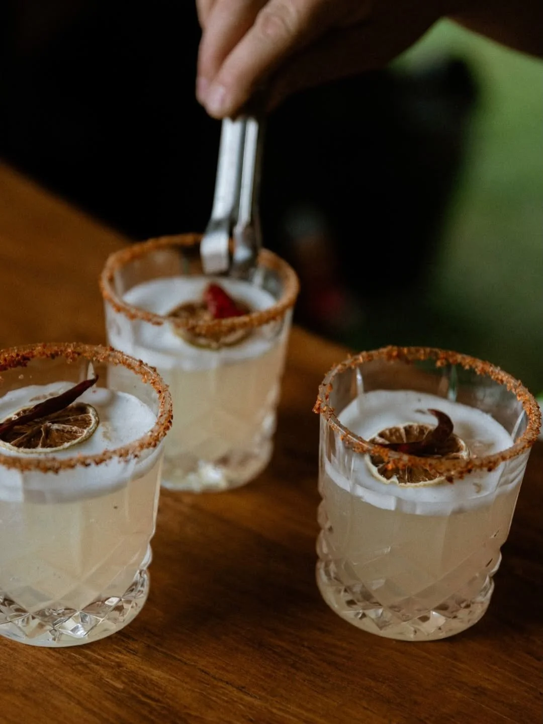 Three glasses of a creamy beverage with a salted rim and dried lime and chili garnishes on top, on a wooden surface, with a hand holding ice tongs above one glass.