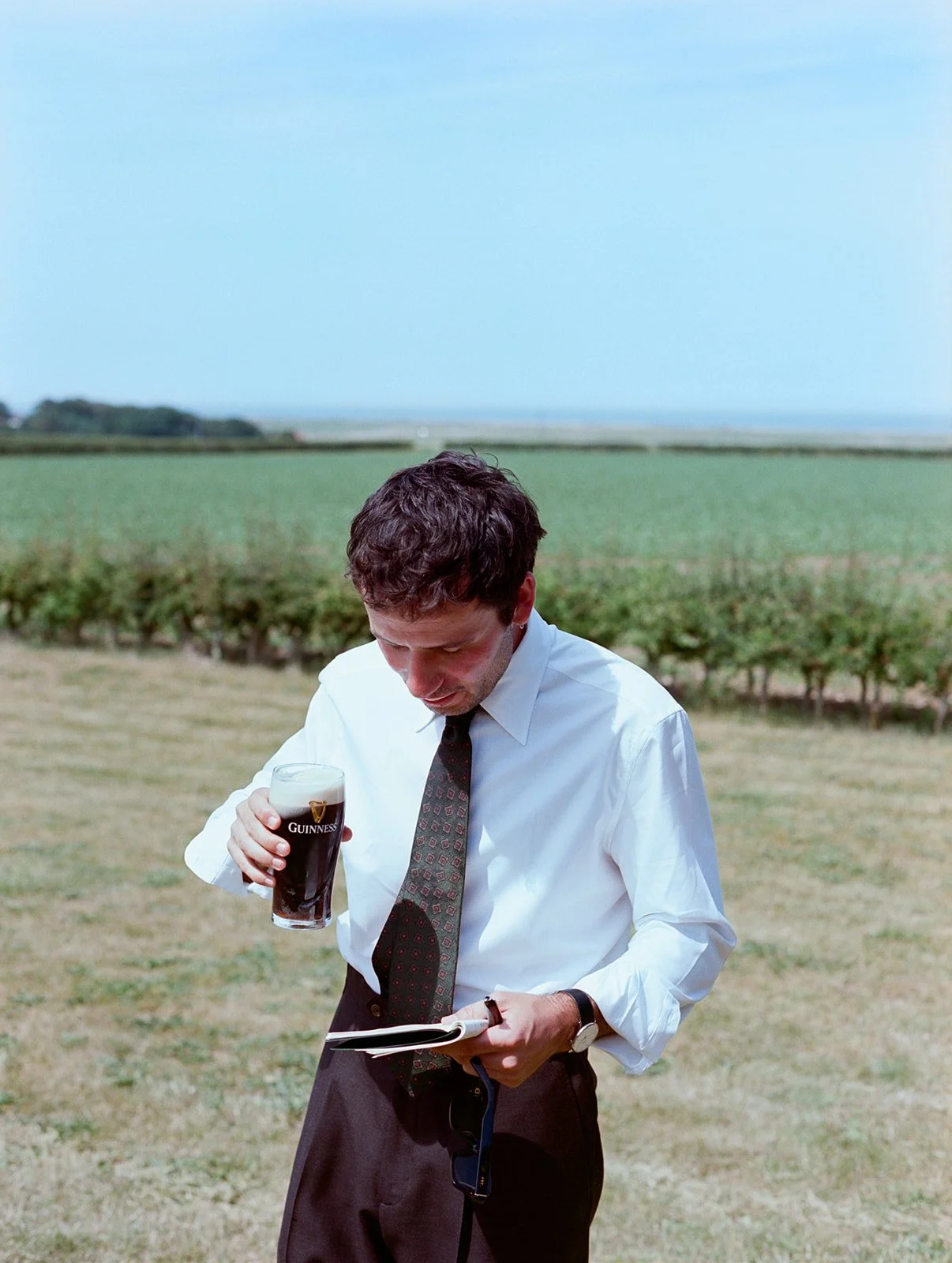 A man in business attire holding a Guinness beer in a field with green crops and an open sky.