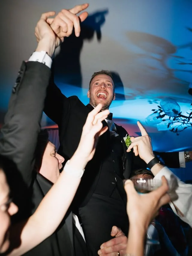 Man in a black tuxedo giving a toast at a celebration, surrounded by people raising their glasses.