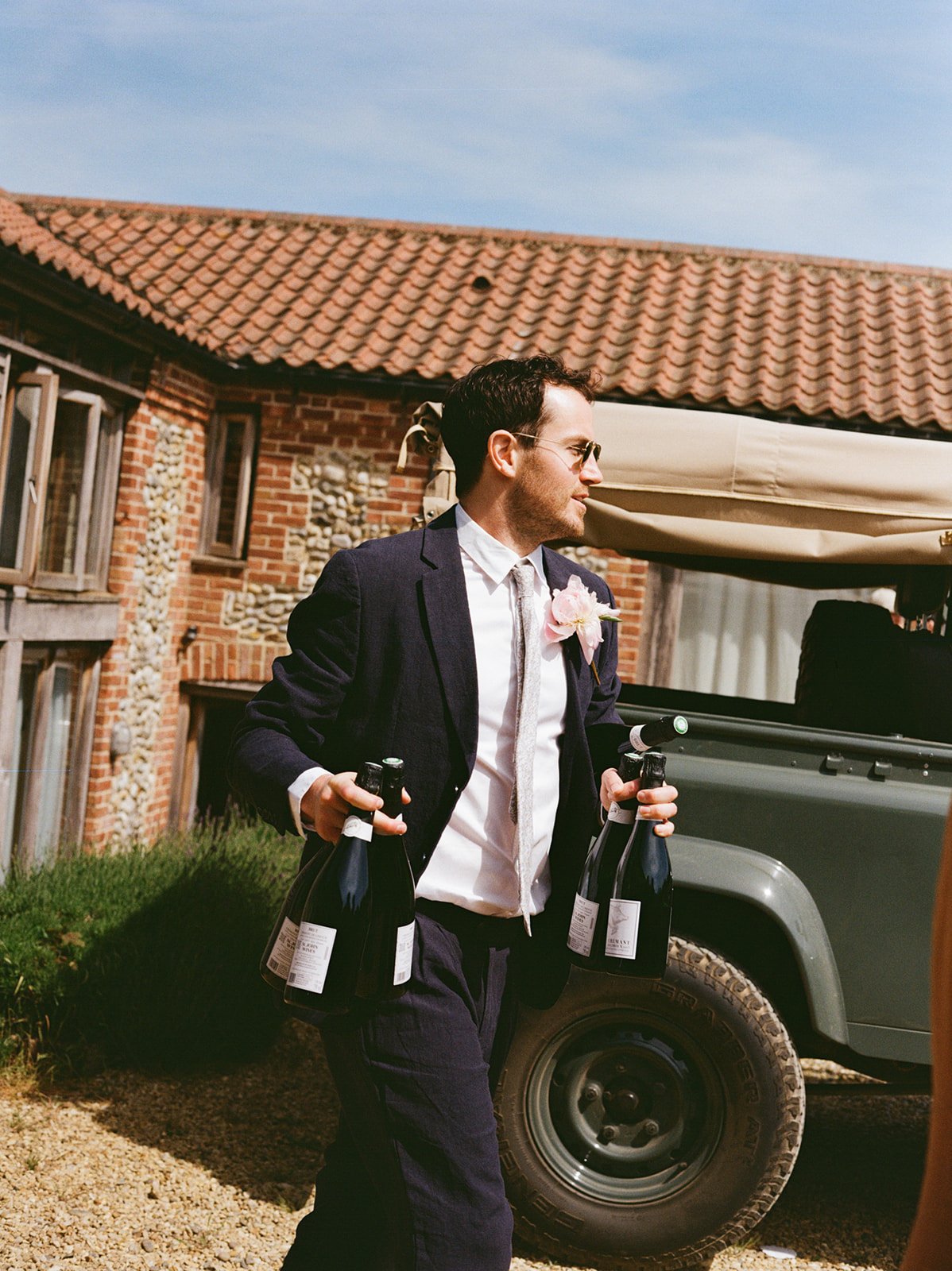 A man wearing a dark suit and tie, with a pink boutonniere, holding two bottles of champagne, standing near a vehicle outdoors in front of a brick house with a tiled roof.