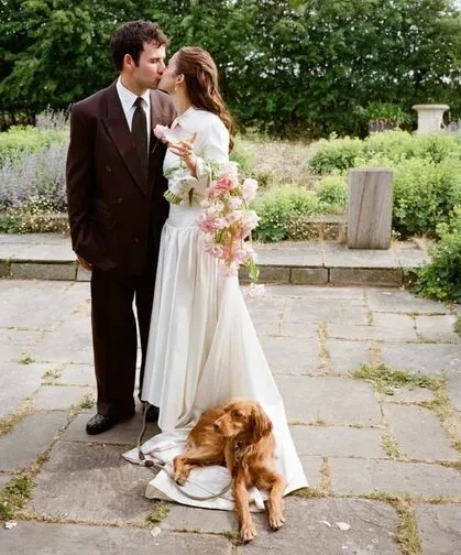 A newlywed couple kissing outdoors on a stone pathway with a dog sitting on the bride's wedding dress, holding a bouquet of pink and white flowers, surrounded by greenery.