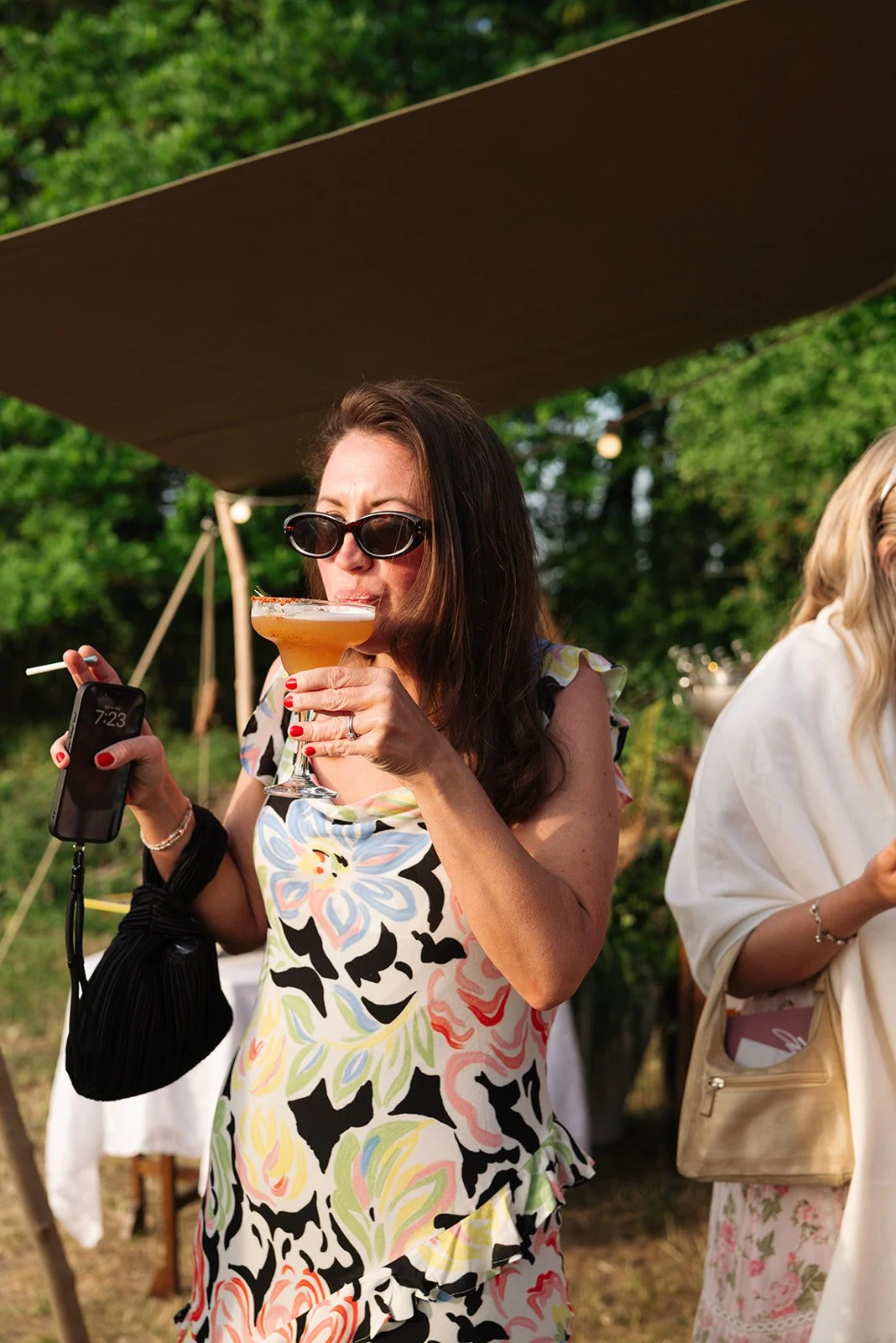 Woman with dark hair, sunglasses, and floral dress sipping a cocktail at an outdoor gathering.