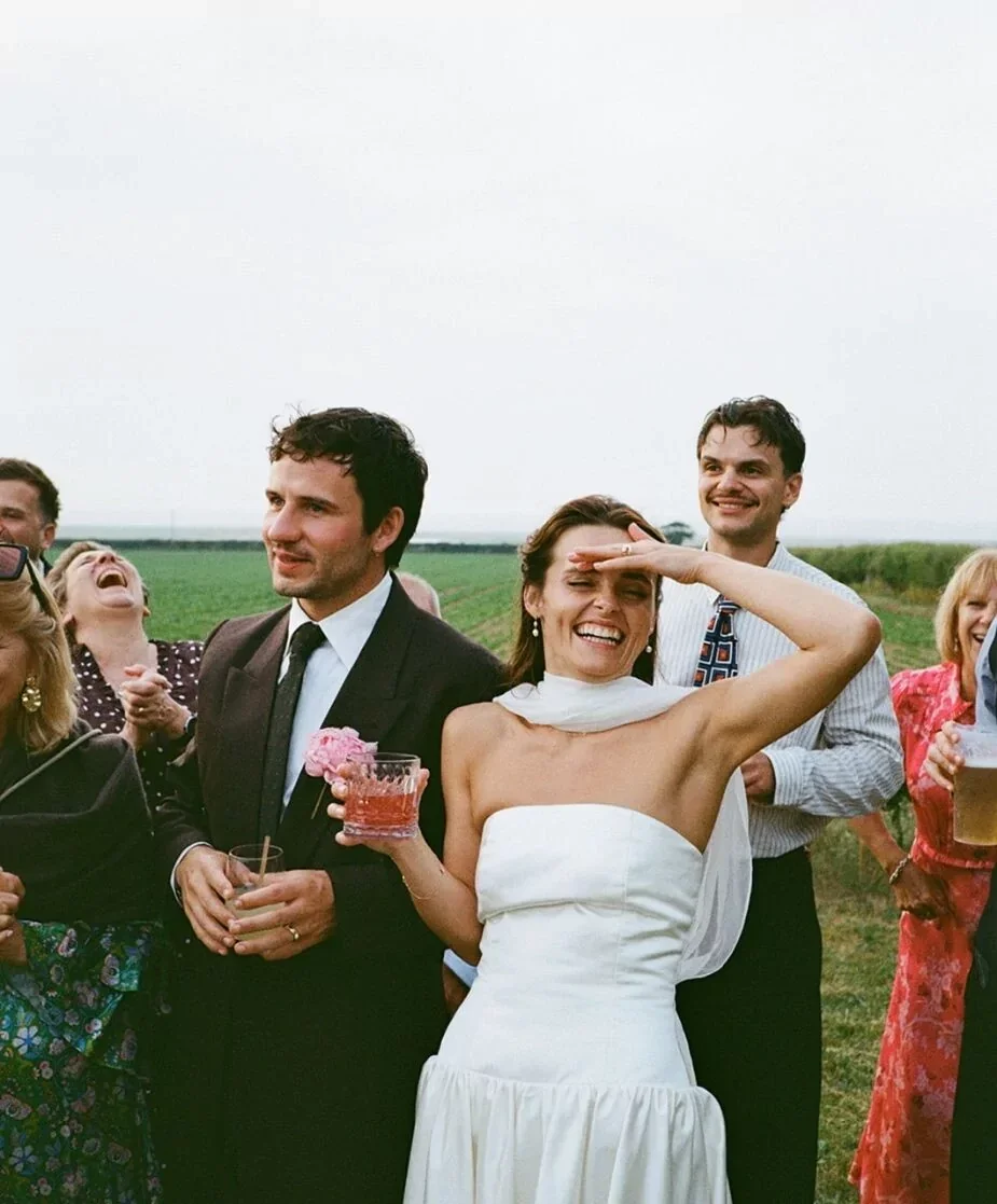 Group of people celebrating outdoors, with a woman in a white dress smiling and shielding her eyes, holding a drink, surrounding a man in a suit, with others laughing and holding drinks.