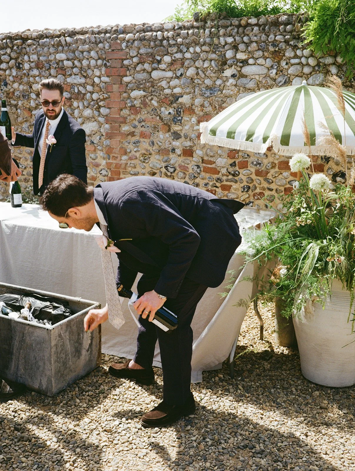 Two men in suits, one bending to pick something from a trash bin, the other standing near a table, outdoors with a stone wall and a green umbrella, in a garden setting with potted plants.