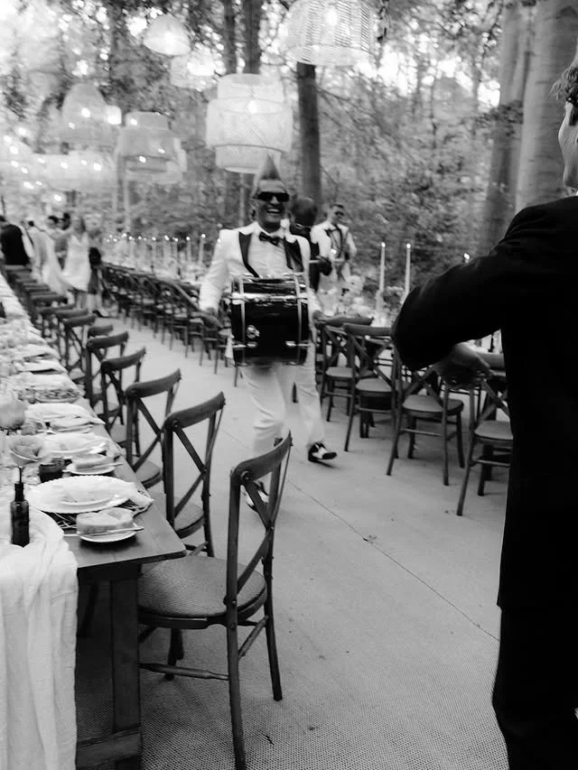 A musician playing drums outdoors, dressed in a white suit with a black bow tie, standing among tables set for an outdoor event or celebration surrounded by trees.
