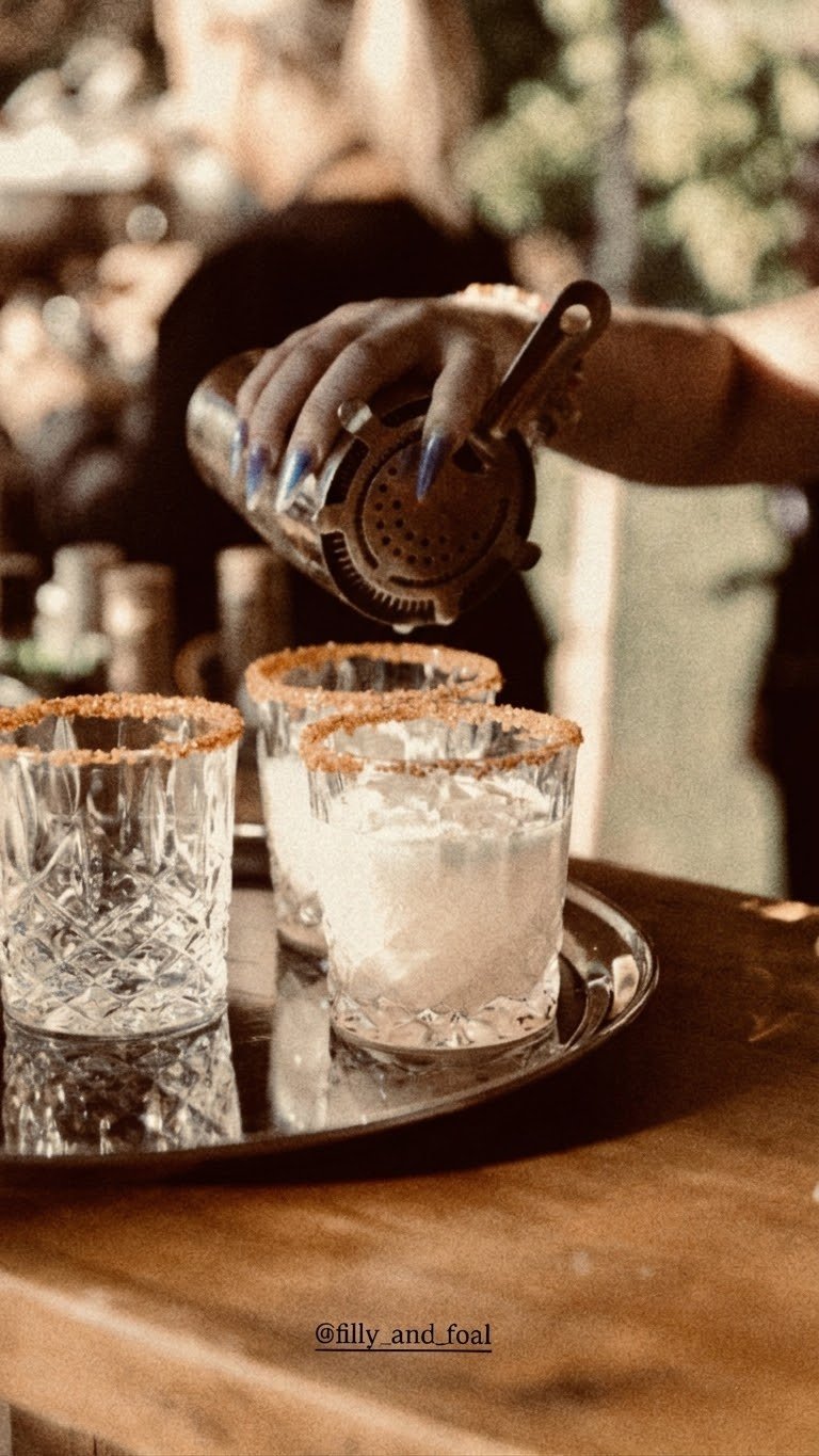 Bartender pouring a frothy cocktail into sugar-rimmed glasses on a mirrored tray