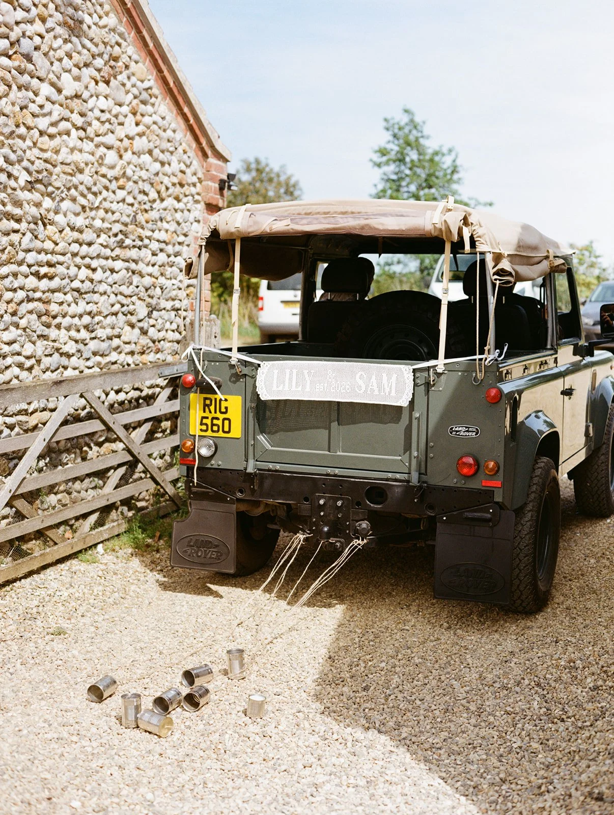 A green Land Rover Defender parked outdoors with a beige soft top. There is a sign on the back that reads 'LILY' and a yellow license plate with 'RIG 560.' The area around the vehicle is covered in gravel, and some empty tin cans are scattered on the ground near the rear. A stone wall and a wooden gate are visible on the left side of the image.
