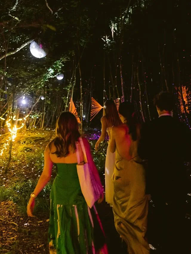 People walking through a dark, wooded area decorated with hanging lanterns and illuminated decorations, likely at an outdoor event or celebration.
