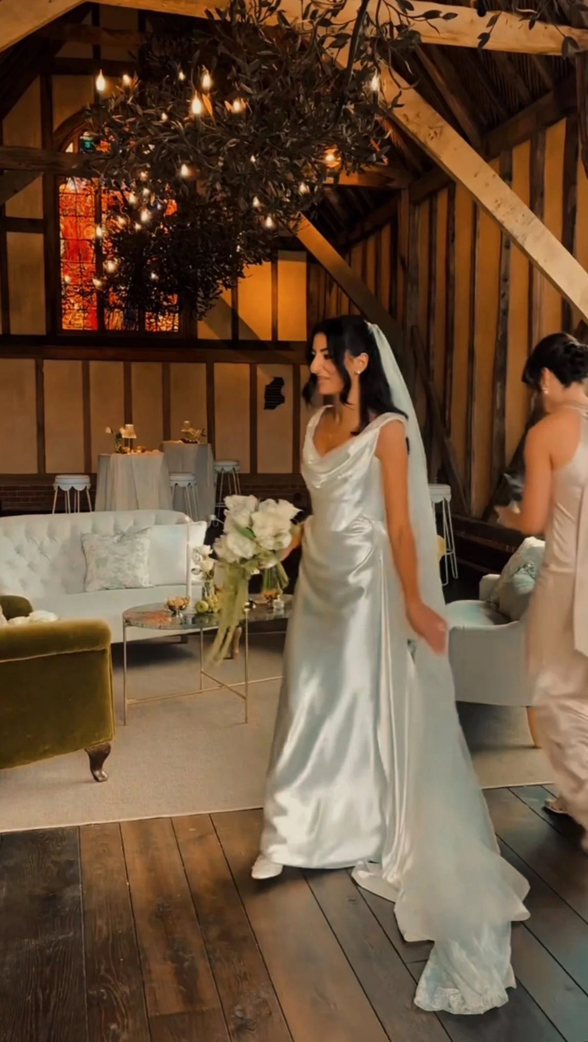 A woman in a white wedding gown holding a bouquet of white flowers, standing in a decorated rustic event space with wooden beams, a chandelier, and elegant furniture.