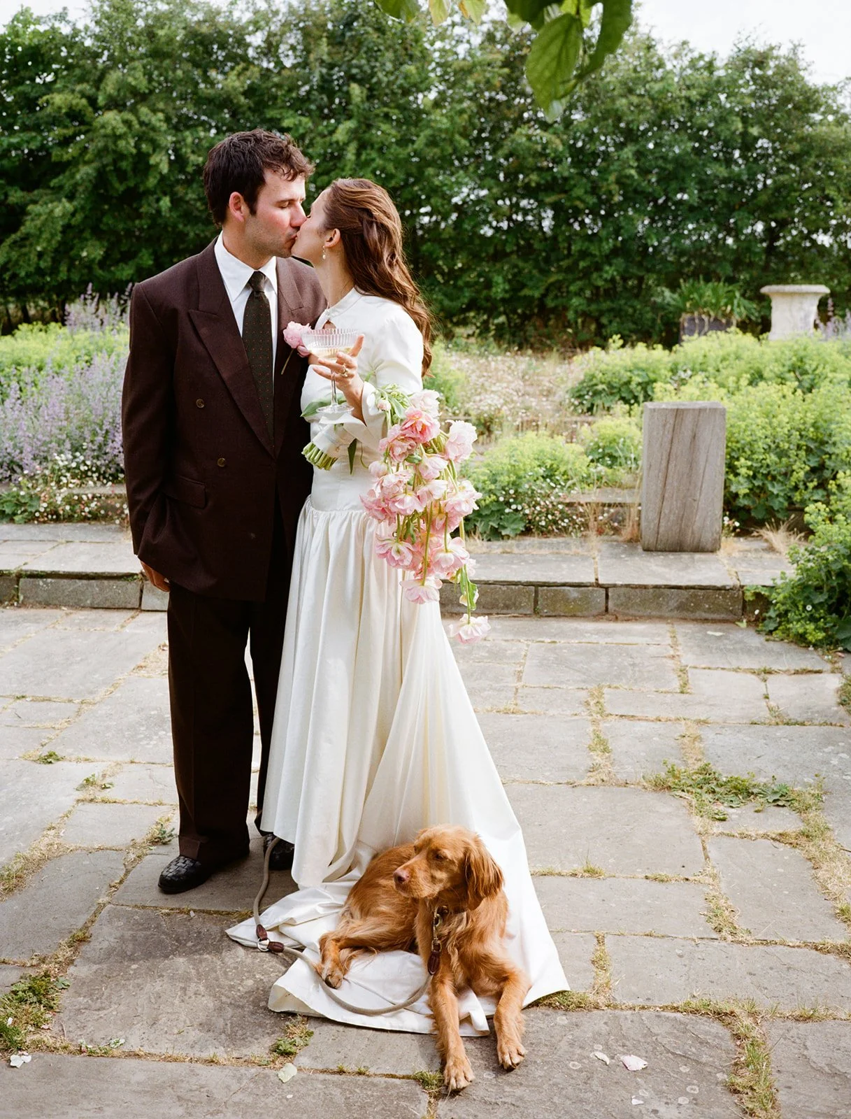 A bride and groom sharing a kiss outdoors in a garden setting, with a dog lying on the ground near the bride's dress and a bouquet of pink flowers.