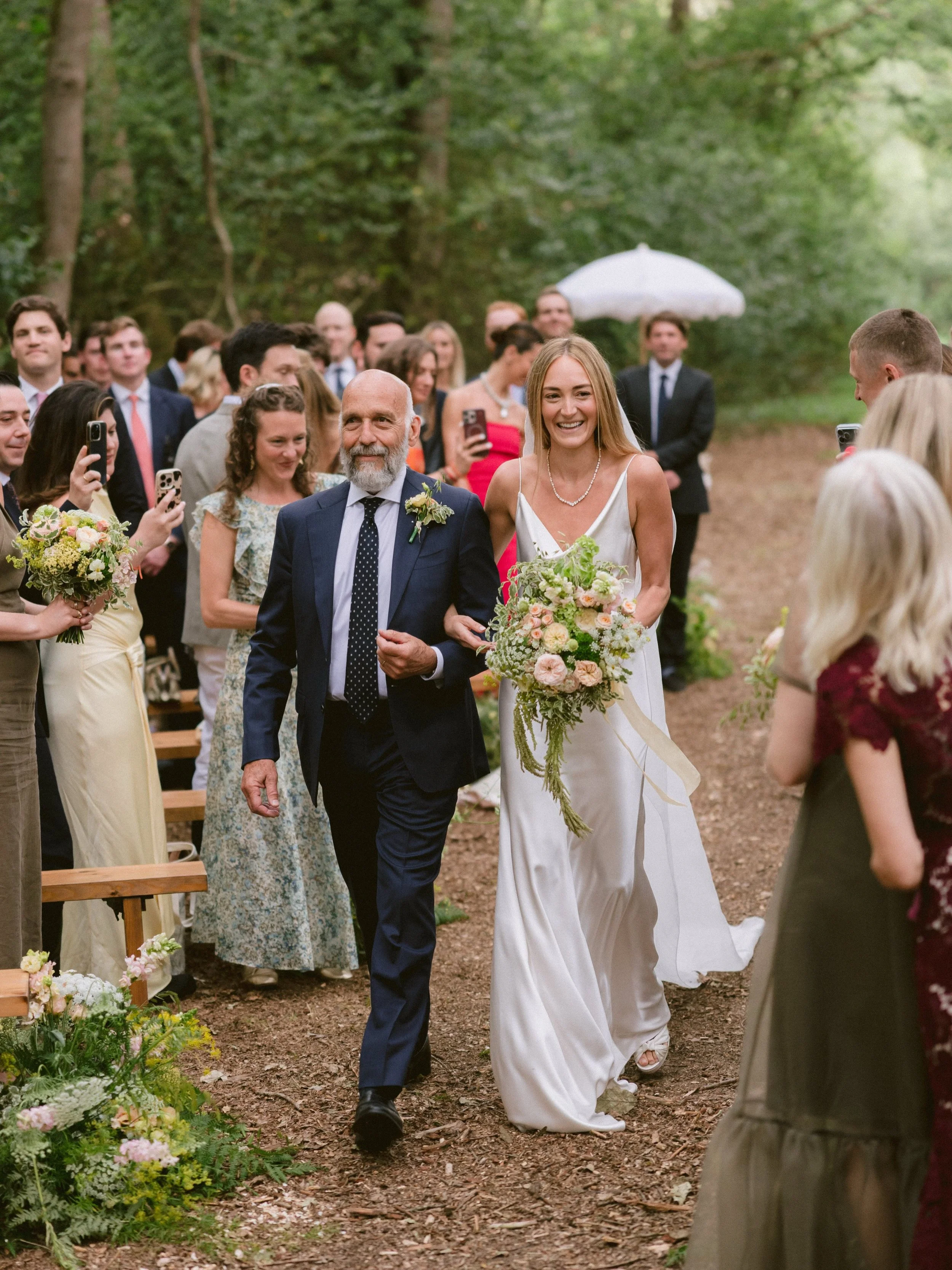 A bride walking arm-in-arm with an older man, likely her father, during an outdoor wedding ceremony in a wooded area. The bride is smiling and holding a bouquet of flowers, while guests are gathered around, some taking photos.