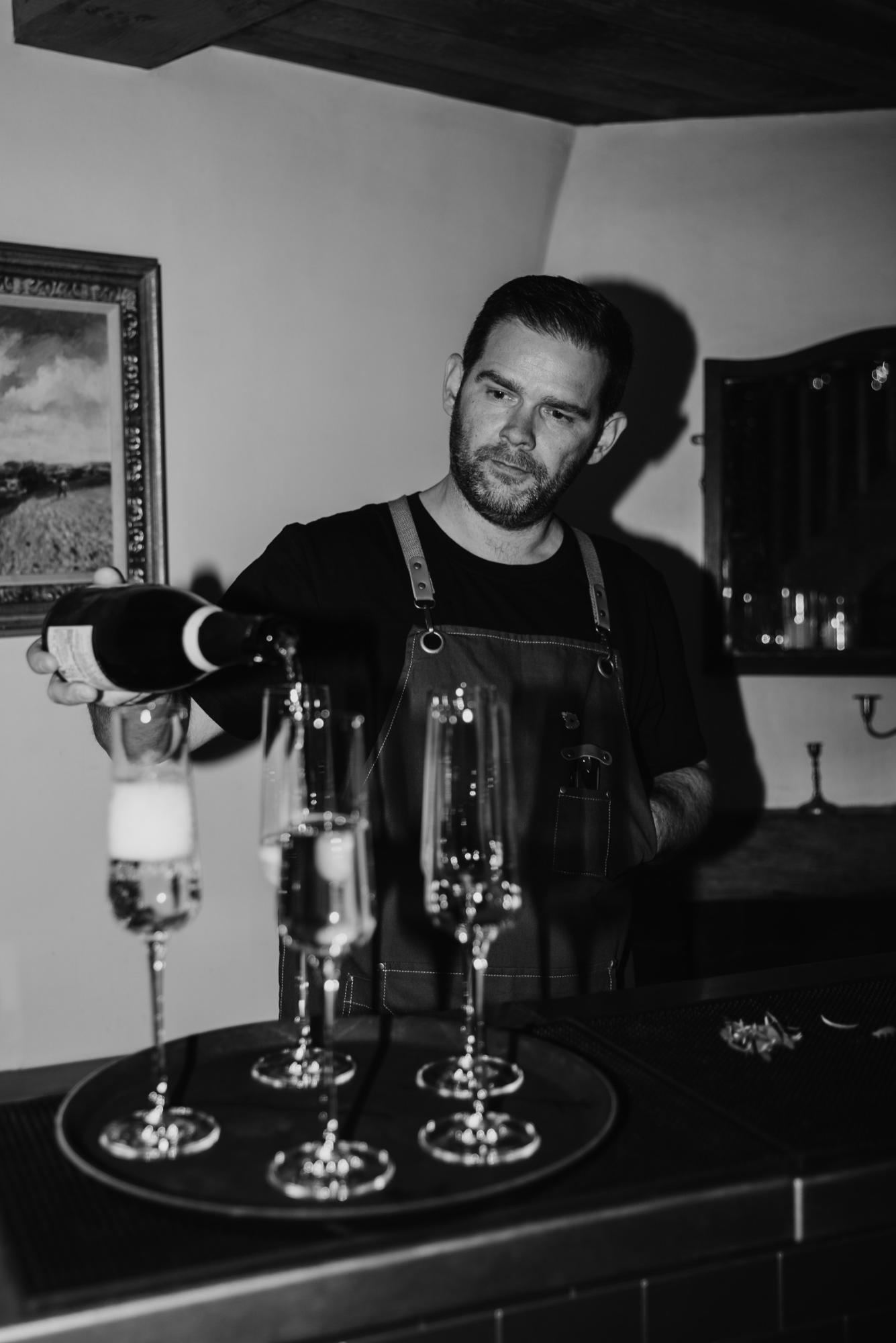 Black and white photo of a man pouring a drink into glasses on a bar counter. The man has dark hair and beard, wearing a dark shirt and apron. In the background, there is a framed picture on the wall and shelves with glasses.