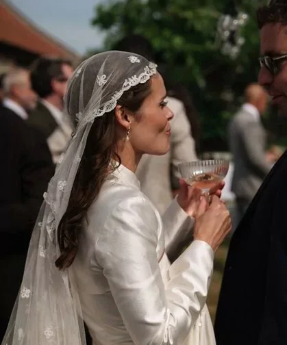 Bride in white wedding dress and veil holding a glass, standing outdoors during a wedding reception, facing a man in dark suit with sunglasses, with other guests in the background.