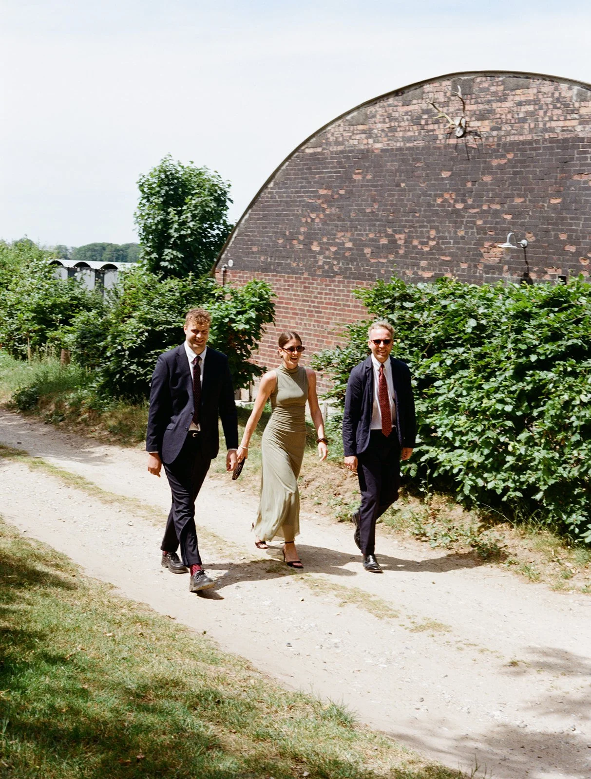 Three adults walking along a dirt path outdoors, with greenery and an arched brick building in the background. The group includes two men in suits and a woman in a sleeveless dress, all smiling.