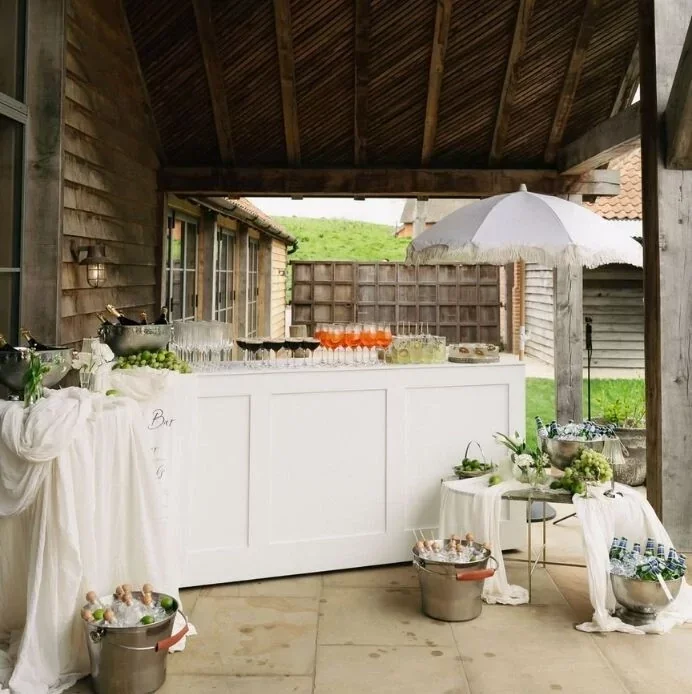 Party drink station under a wooden pavilion with a white bar, an umbrella, and various beverages and snacks on display.