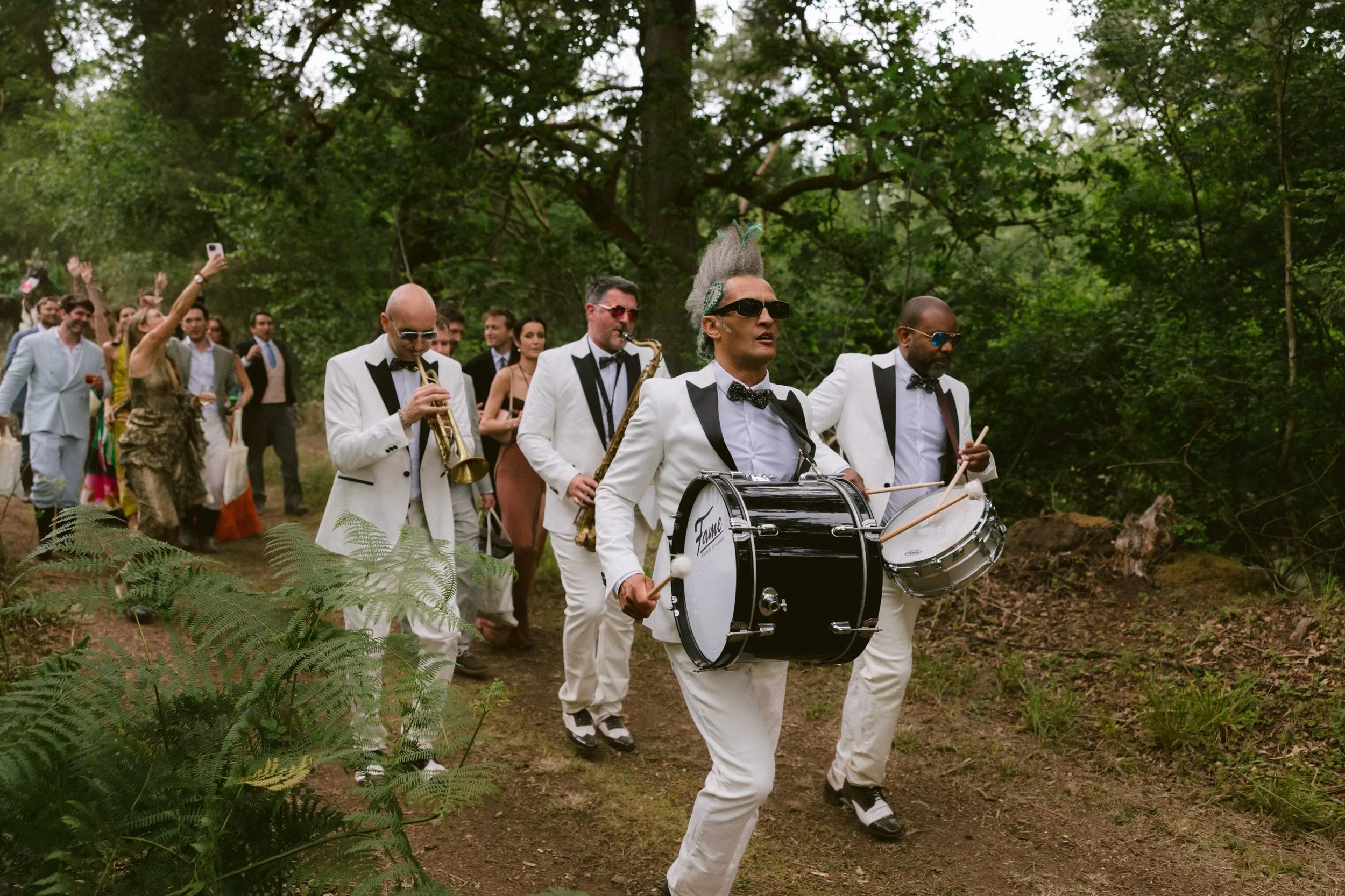 A group of people in formal attire, including men in tuxedos with bow ties and women in dresses, walking through a wooded area. A man in the forefront is playing a large bass drum, and other musicians are playing wind instruments. The group appears to be part of a parade or celebration on a forested path.