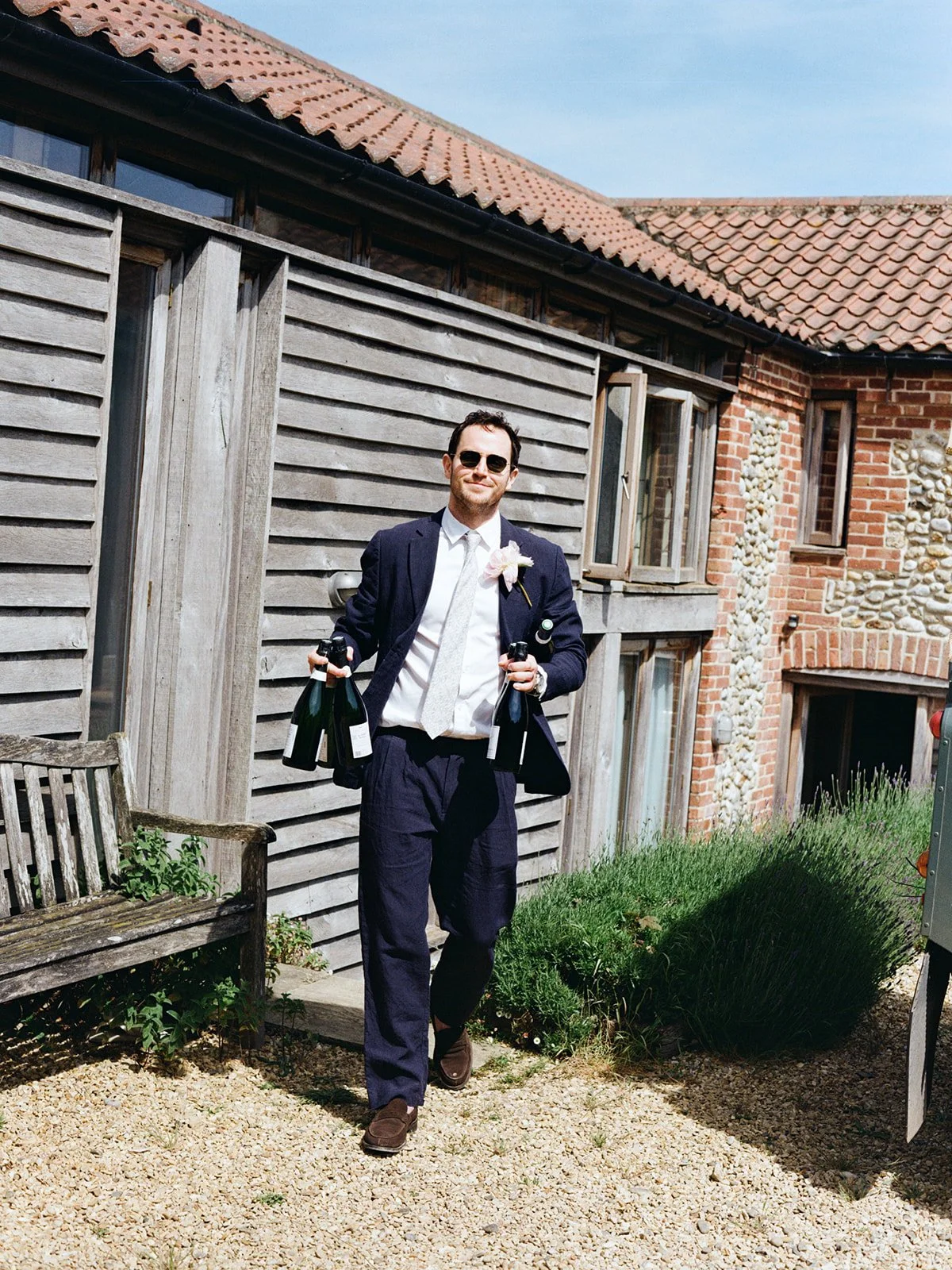 A man dressed in a suit with a boutonniere, sunglasses, and tie, holding bottles of champagne in each hand, walking outdoors near a building with a wooden and brick exterior, under a blue sky.