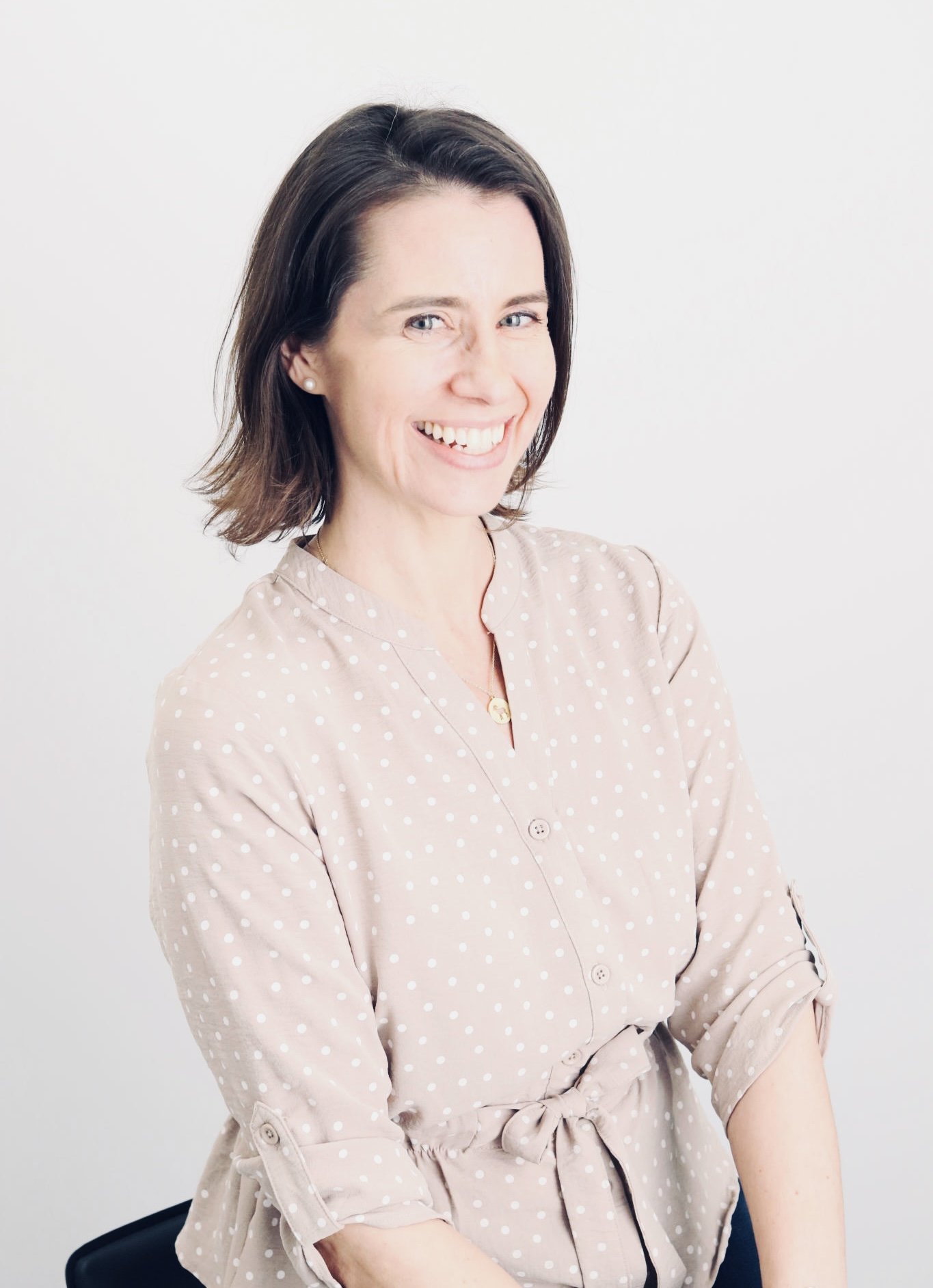 Smiling woman with short brown hair wearing a pale pink polka-dot dress against a white background