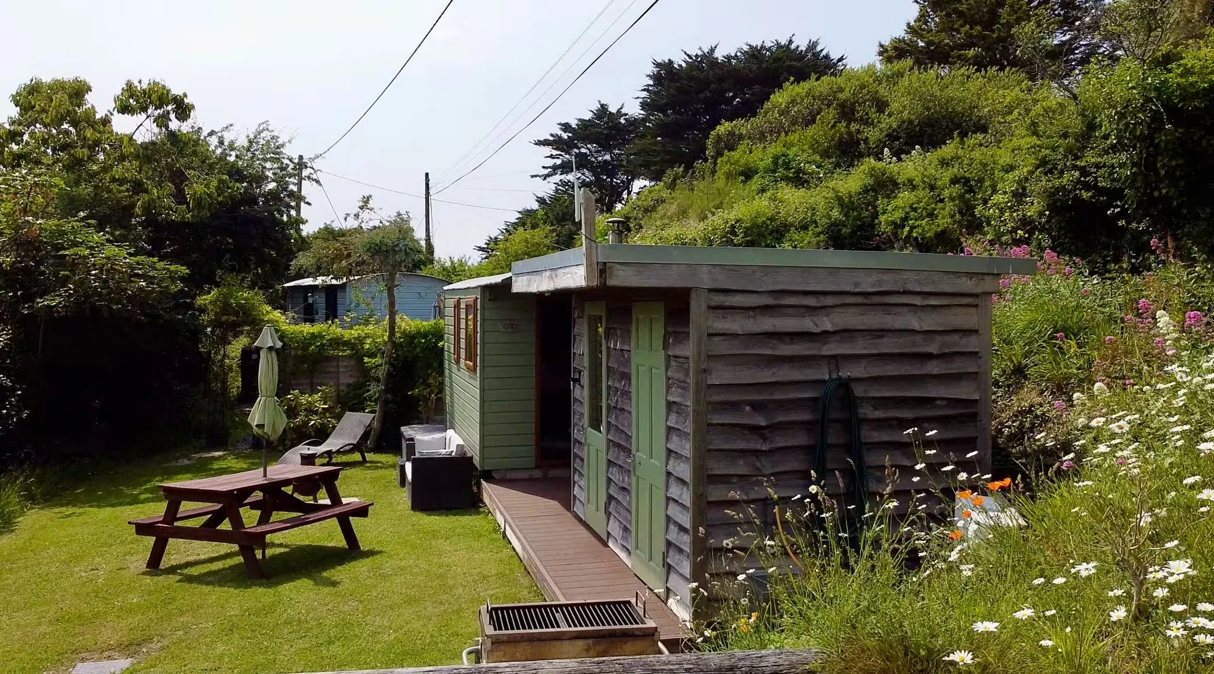 View of Foxes Cove and garden area, surrounded by trees, flowers, and garden furniture including a picnic table and lounge chairs.