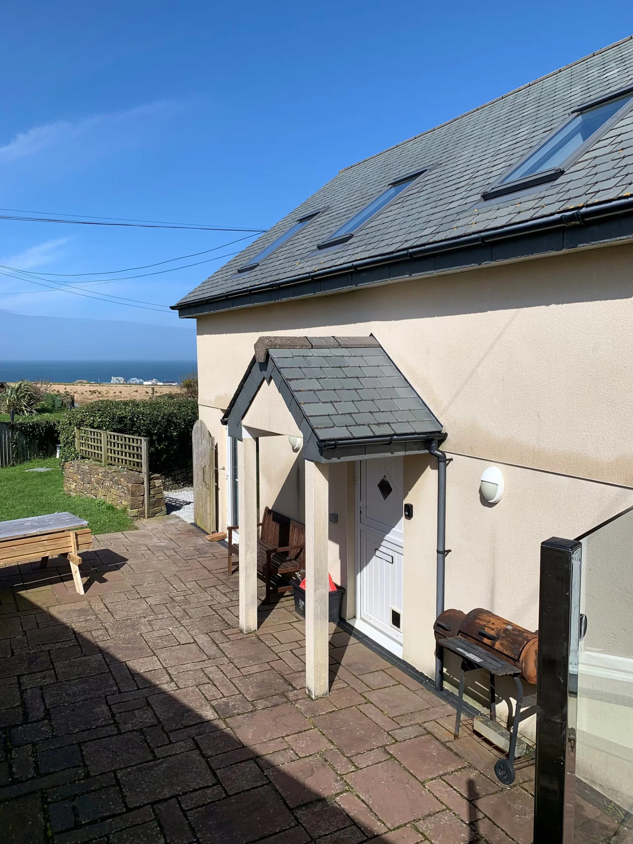 Backgarden patio with brick paving, wooden benches, a white house entrance with a small porch, a barbecue grill, and a view of the ocean in the background under a clear blue sky.