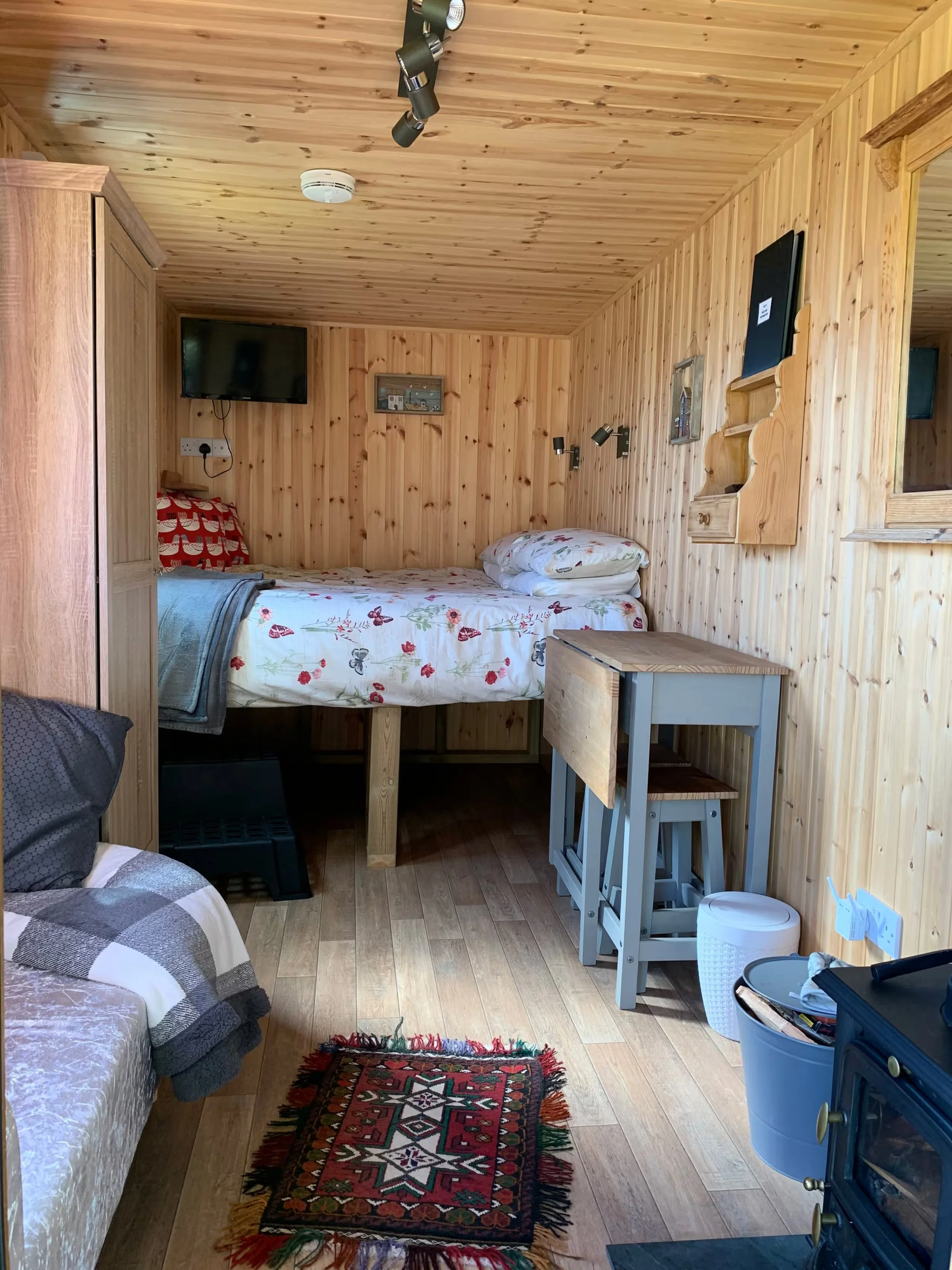 A cozy wooden-paneled bedroom at Foxes Cove featuring a small bed, a patterned rug on the floor, a small wooden table with stools, a wall-mounted TV, and various lighting fixtures.