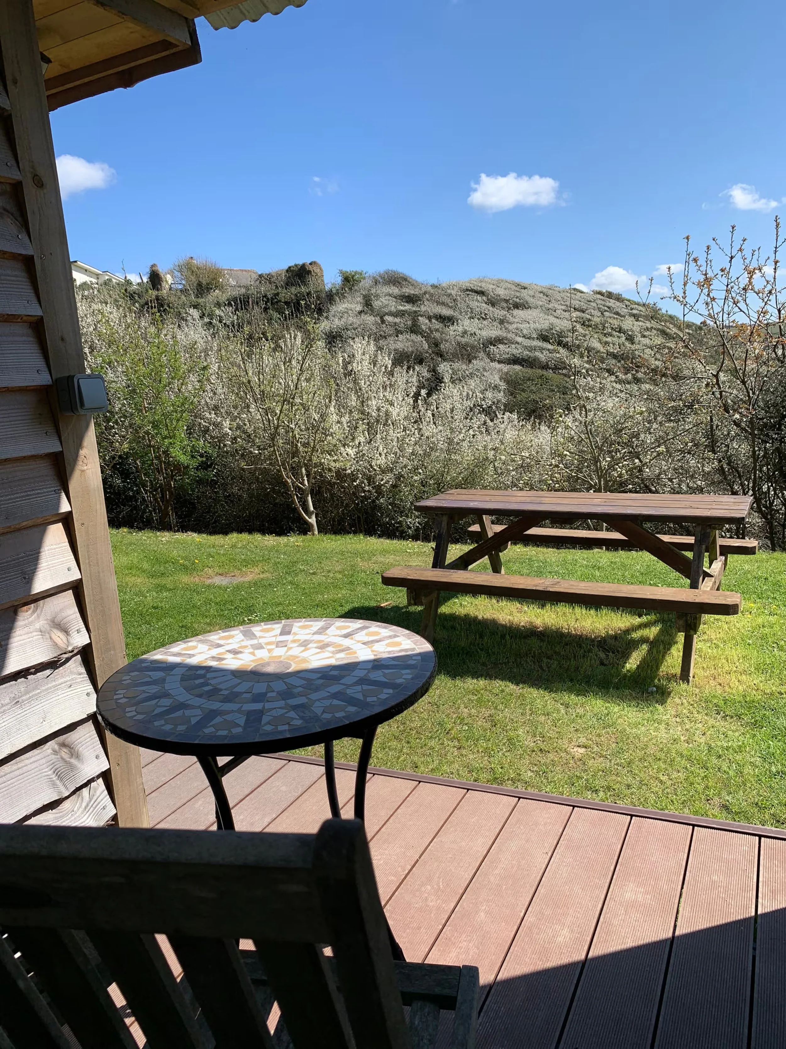 A view from the deck of Foxes Cove with a table, a picnic table, overlooking a landscape with blooming white trees on a hillside under a blue sky with scattered clouds.