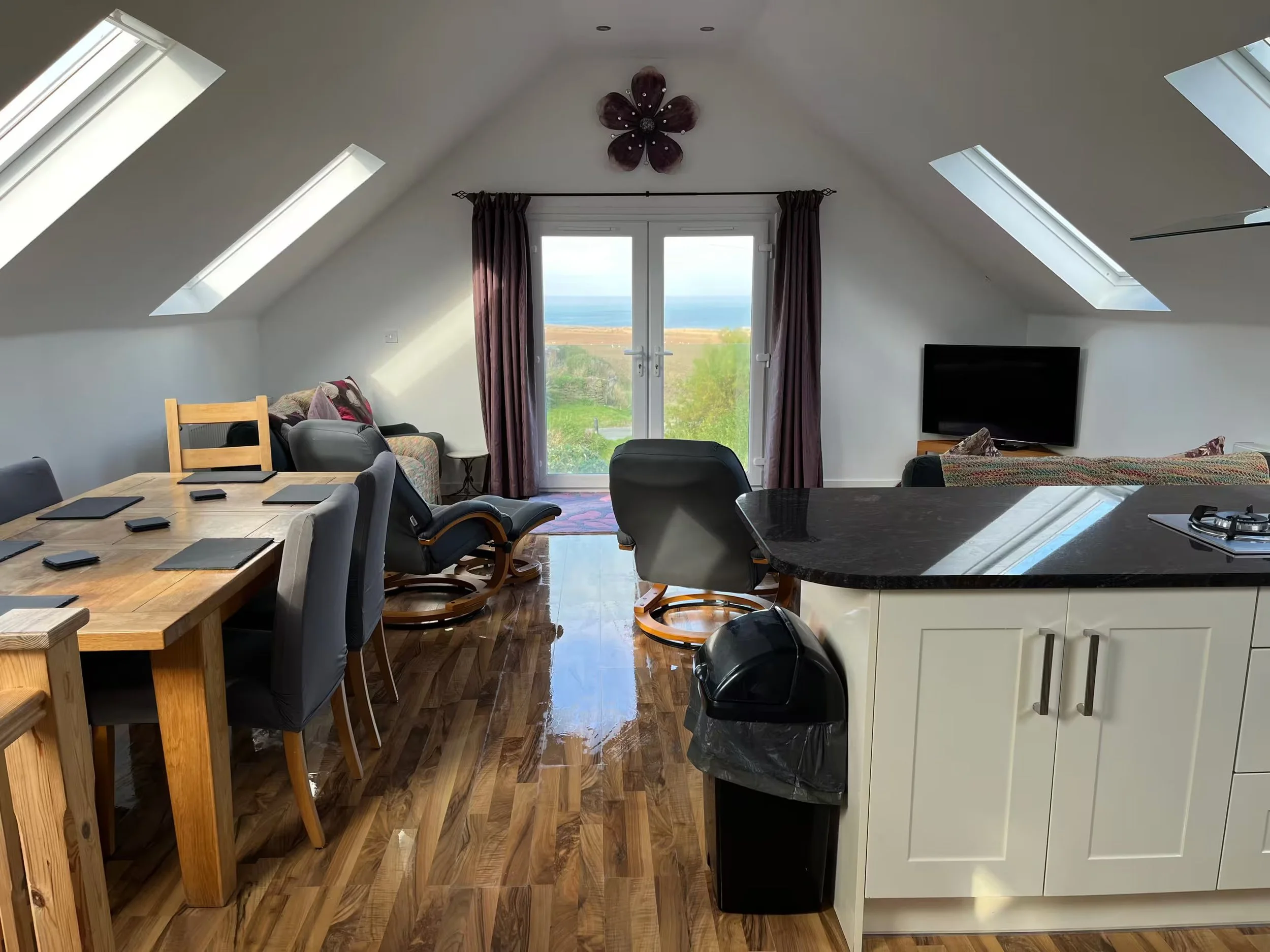 Interior view of living space with a large window and skylights, showing a dining area with a wooden table, chairs, and a kitchen counter with a stove, and a view of the ocean.