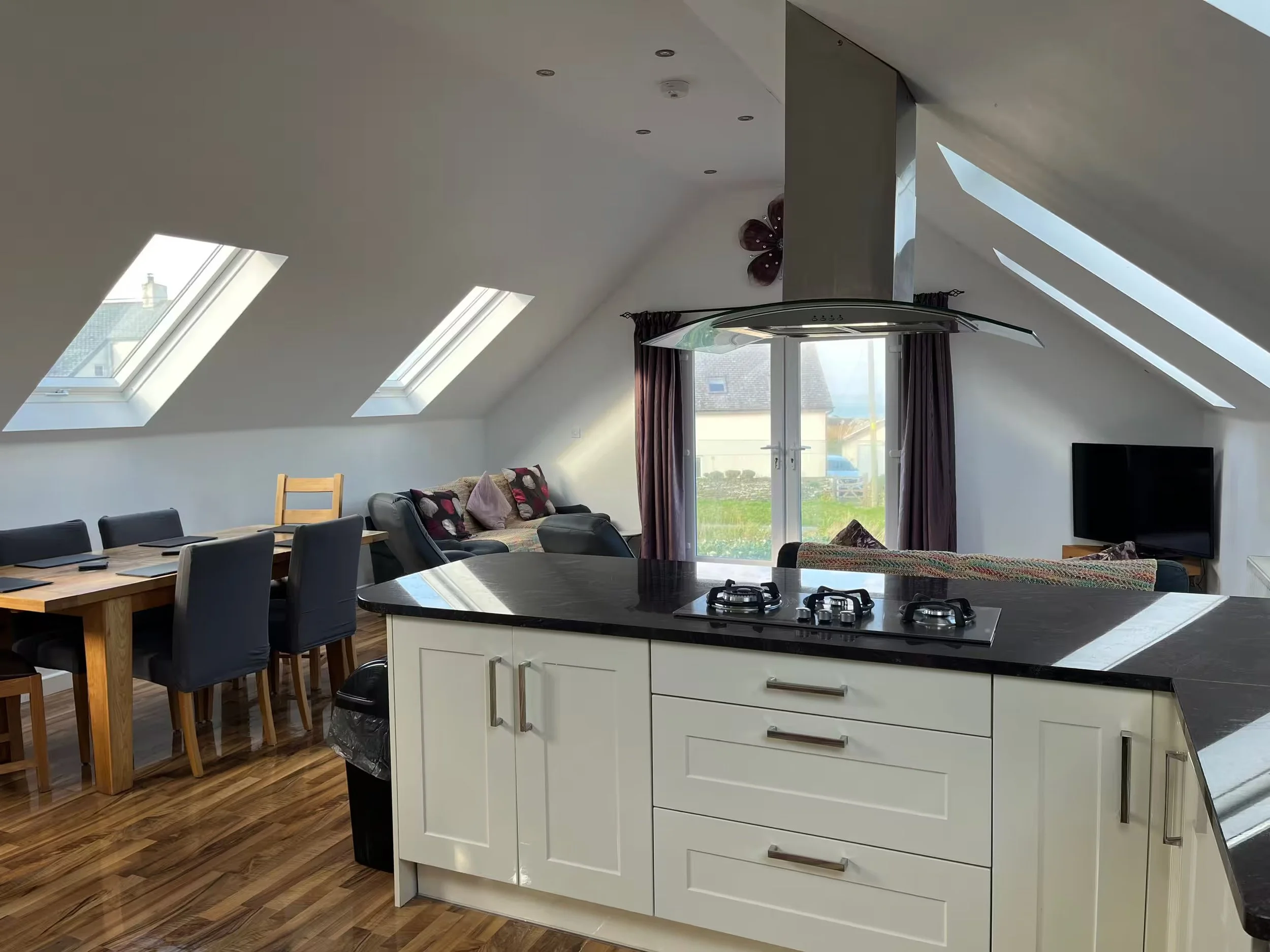 A spacious, modern kitchen with a white island and black countertop, stainless steel appliances, a dining table with black chairs, and large skylight windows letting in natural light. There is a living area with a sofa and curtains visible through a 