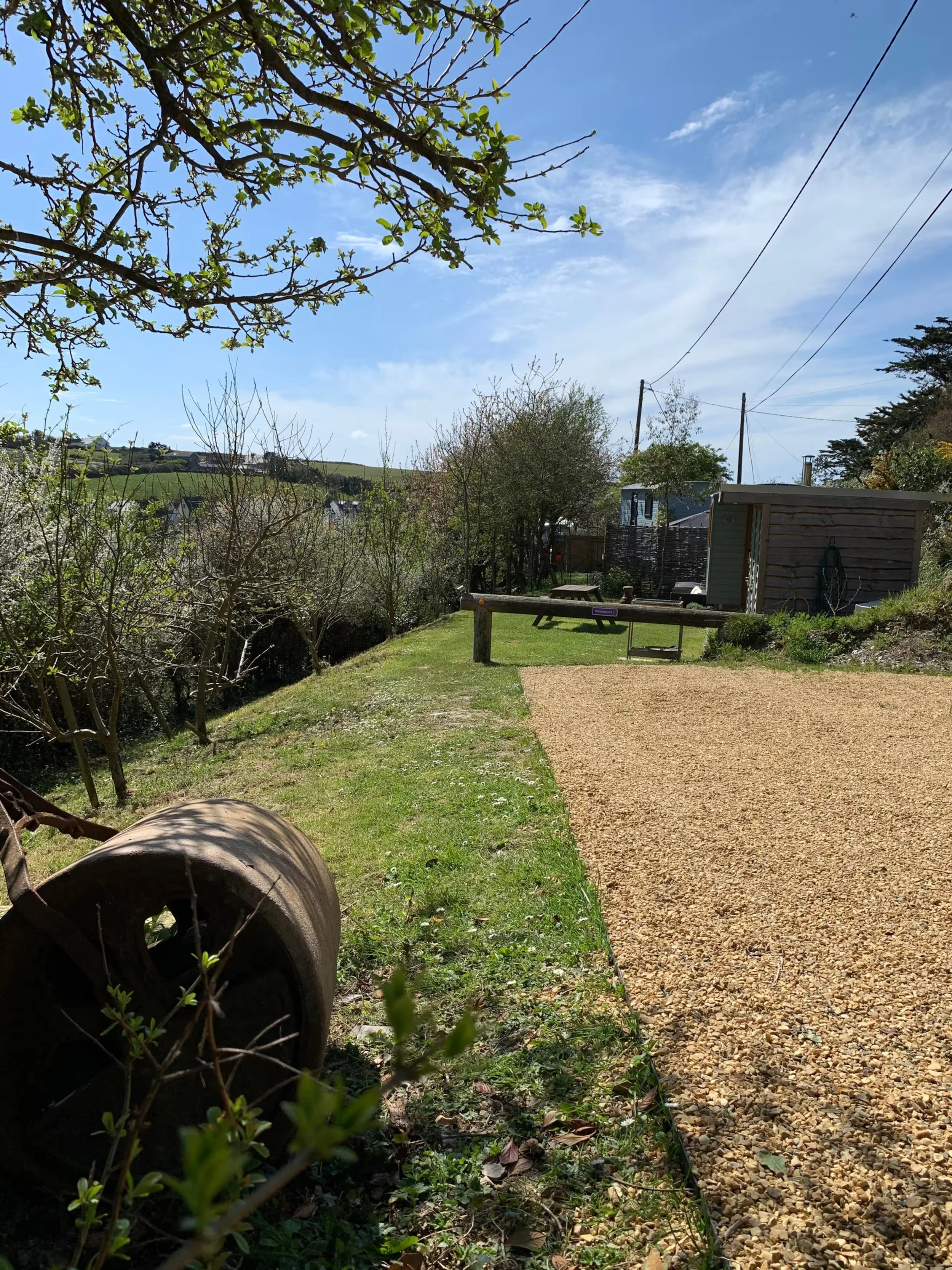 Parking at Porthcothan Retreats with a grassy area, a gravel pathway, trees with budding leaves.