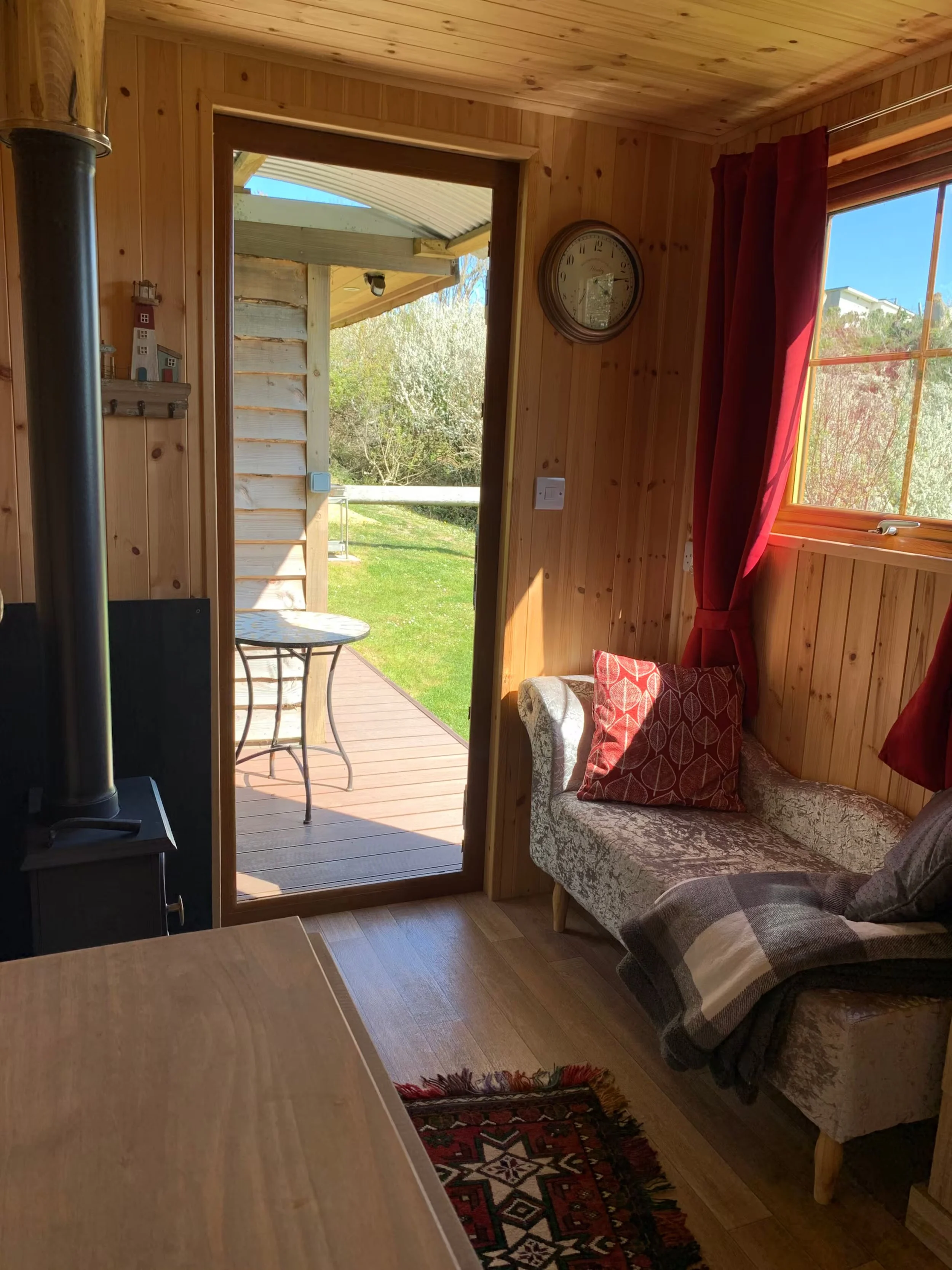 Inside Foxes Cove, a cozy wooden cabin with a doorway leading to a porch with outdoor furniture, a sofa with cushions, a window with curtains, a clock on the wall, and trees outside.
