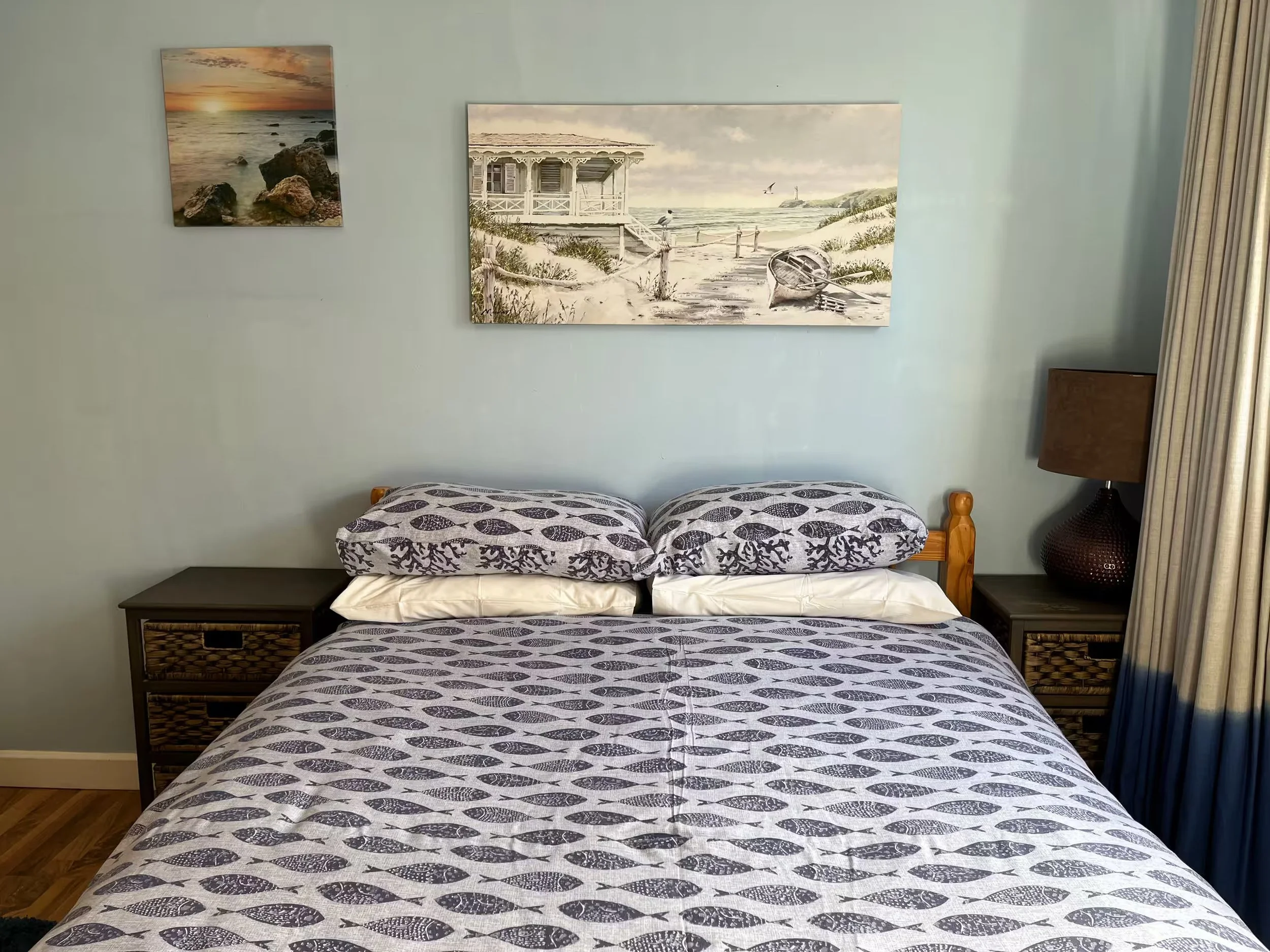 Interior of a bedroom with a bed covered in a black and white fish patterned bedspread, two wicker side tables, framed beach and ocean landscape paintings on a pale green wall, and beige curtains to the side.