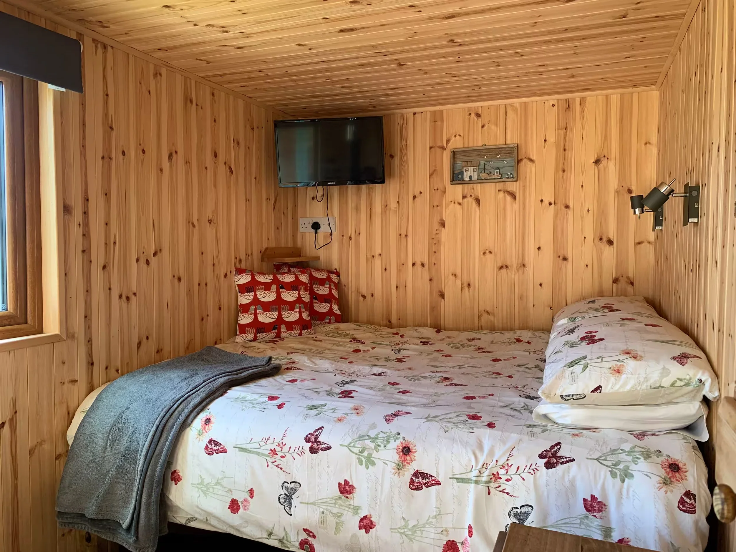 A cozy bedroom with wood-paneled walls and ceiling, featuring a bed with floral and butterfly-patterned bedding, a pillow, a folded gray blanket, a small bedside table, a wall-mounted television, a framed artwork, and a wall-mounted reading lamp.