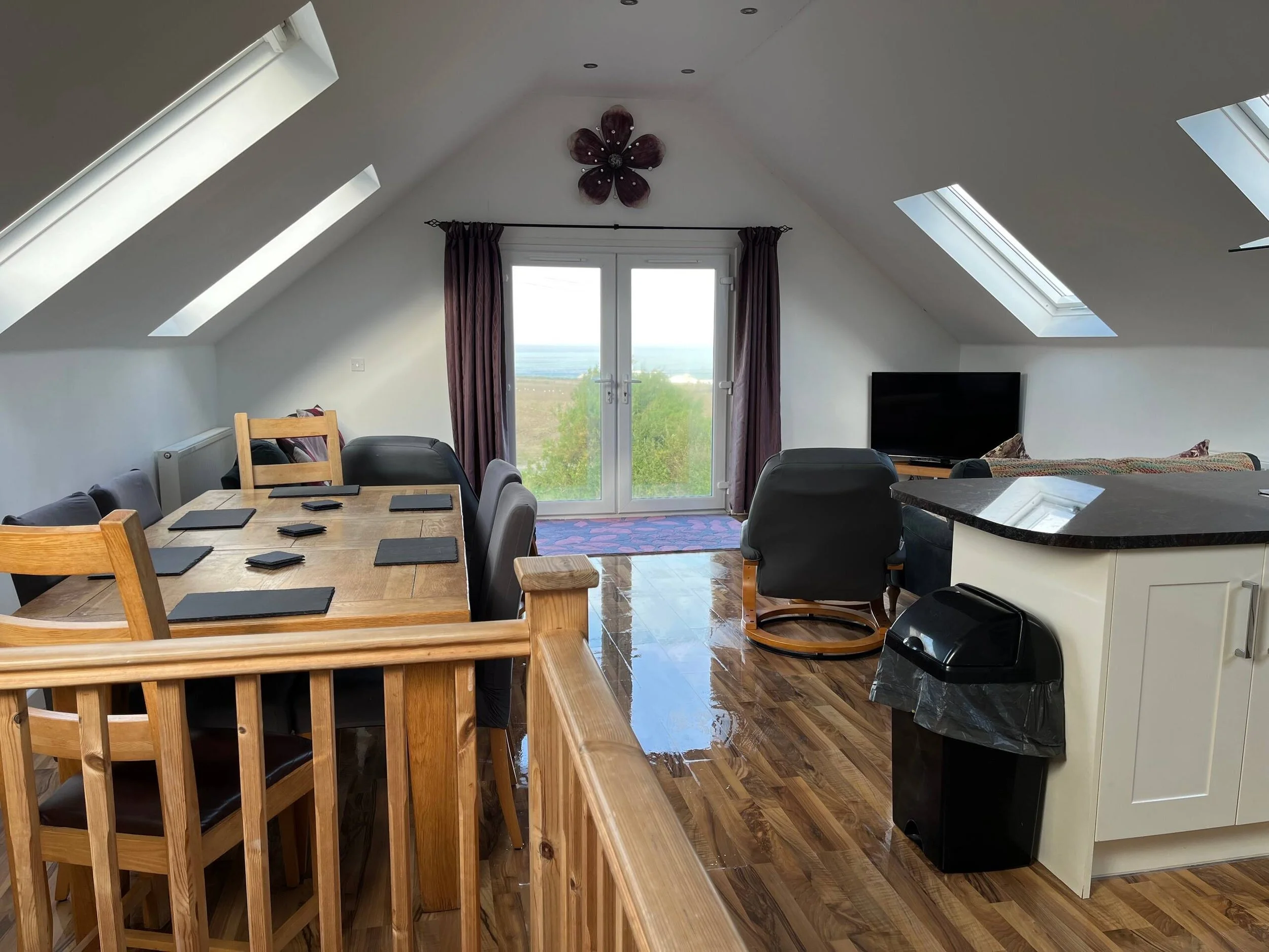 Living room with sloped ceilings and skylights, a sliding glass door with purple curtains, a dining table with black placemats, a black TV, and a modern chandelier shaped like a flower.