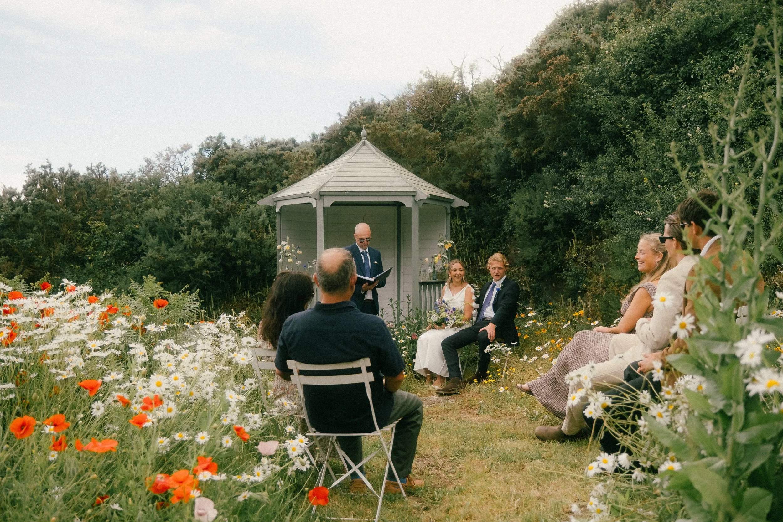 A wedding ceremony outdoors in a garden with flowers and greenery. The bride and groom are seated in front of a small white gazebo, with a man reading vows or a speech. Guests are seated nearby, smiling and watching the ceremony.